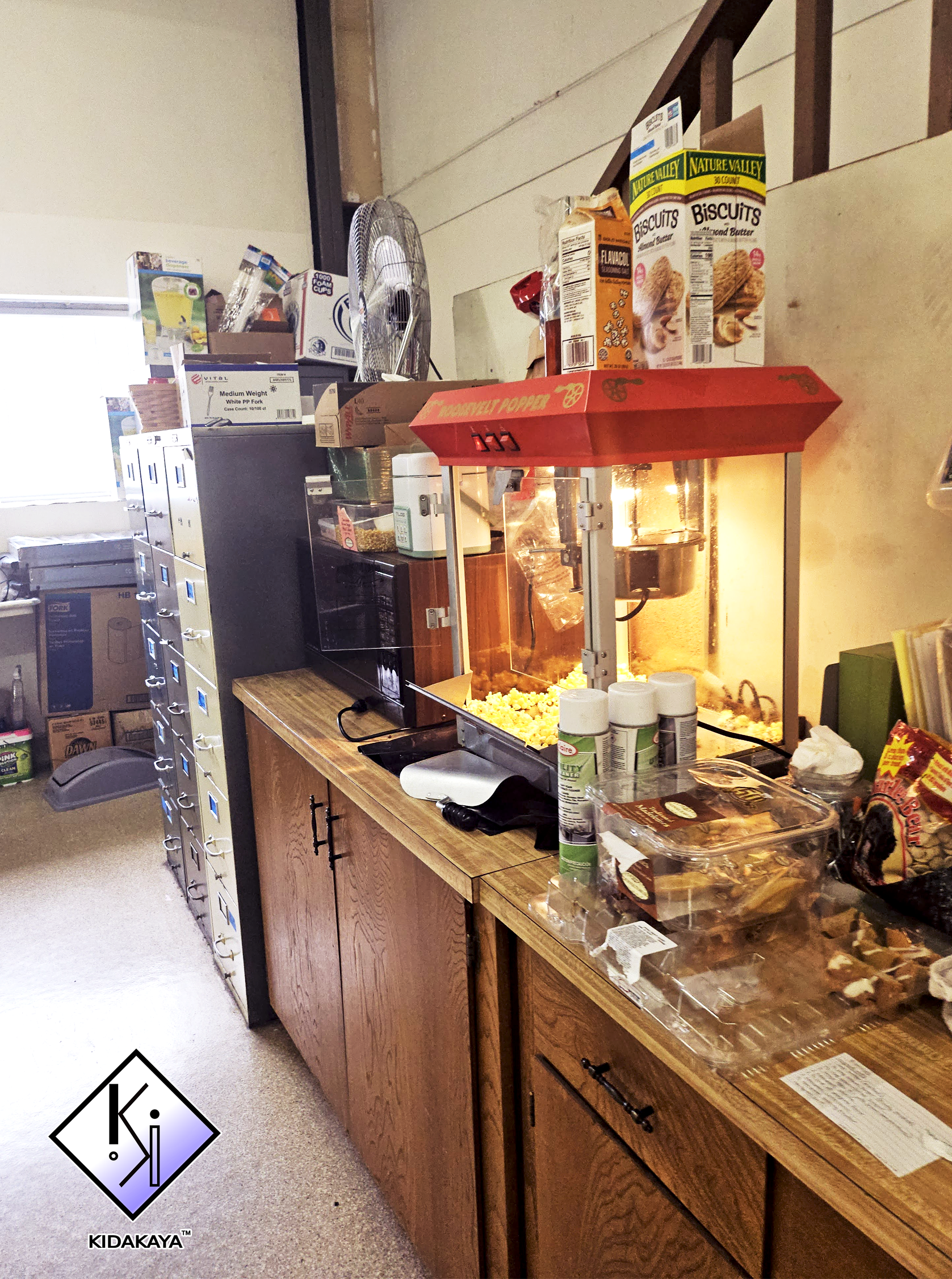 A food service area with a popcorn machine on a wooden counter, snacks, and canned drinks, against a wall with a small window and filing cabinets in the background.