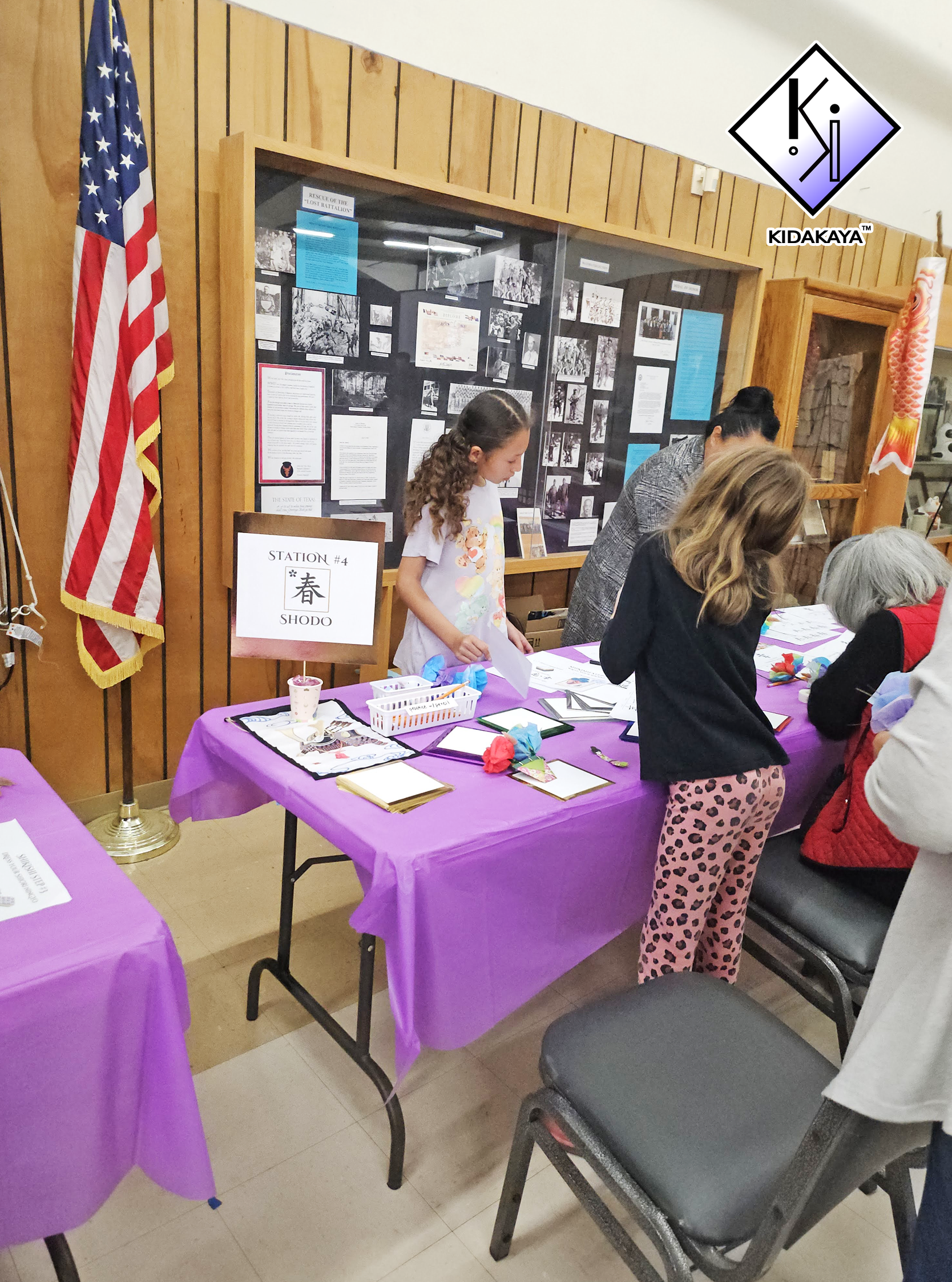 People at a table with purple tablecloths participating in an activity, with informational display on the wall and an American flag nearby.