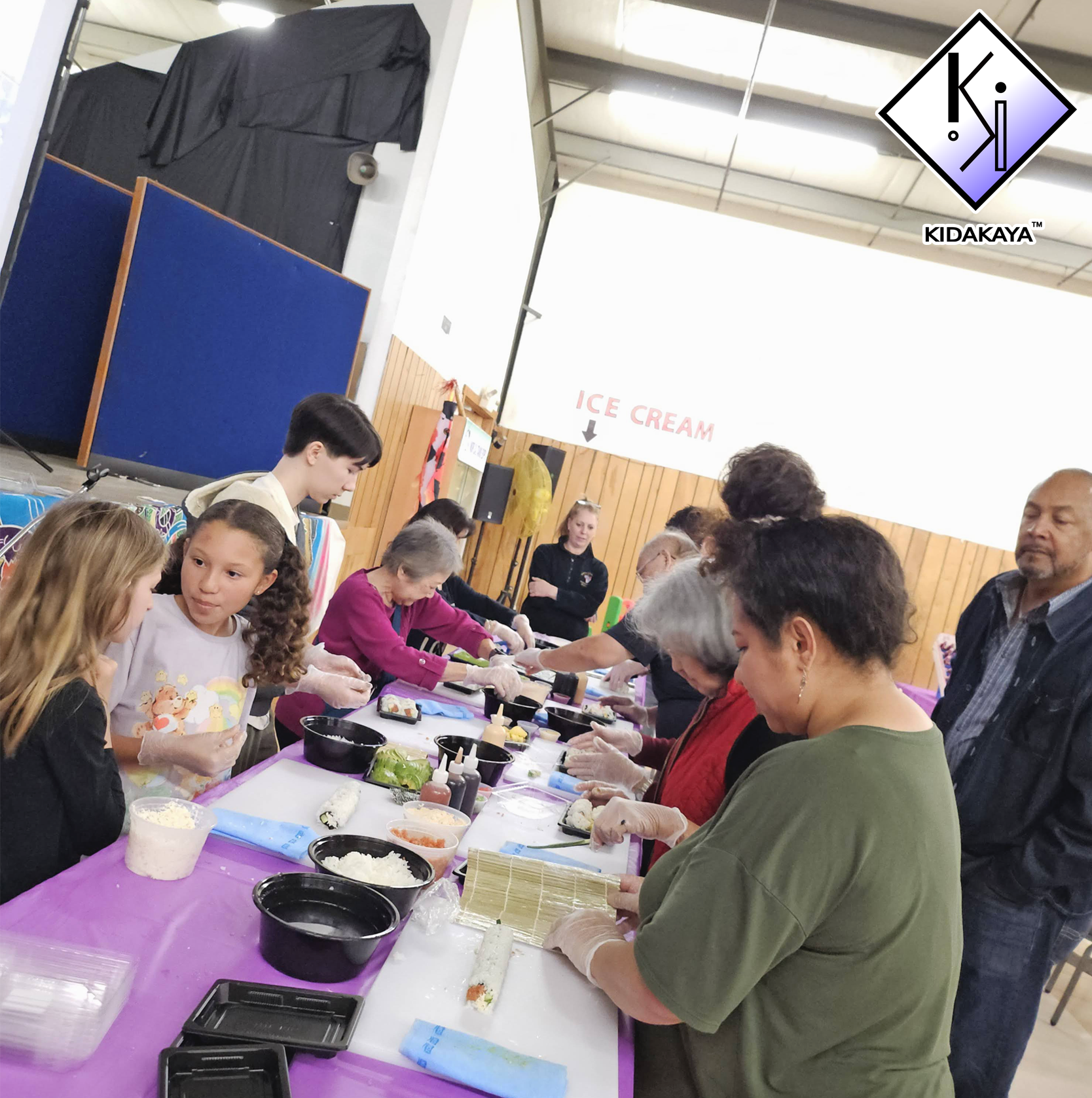 A group of people gathered around a table preparing sushi, with ingredients like rice, seaweed, and vegetables. The setting appears to be a community center or classroom with a wooden wall and a sign saying 'Ice Cream' in the background.