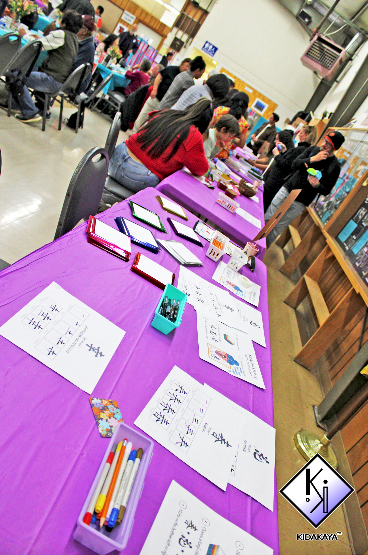 A table covered with a purple tablecloth displaying worksheets, colorful pens, and small stationery items at a cultural event. People are gathered around another table in the background, participating in activities or socializing.