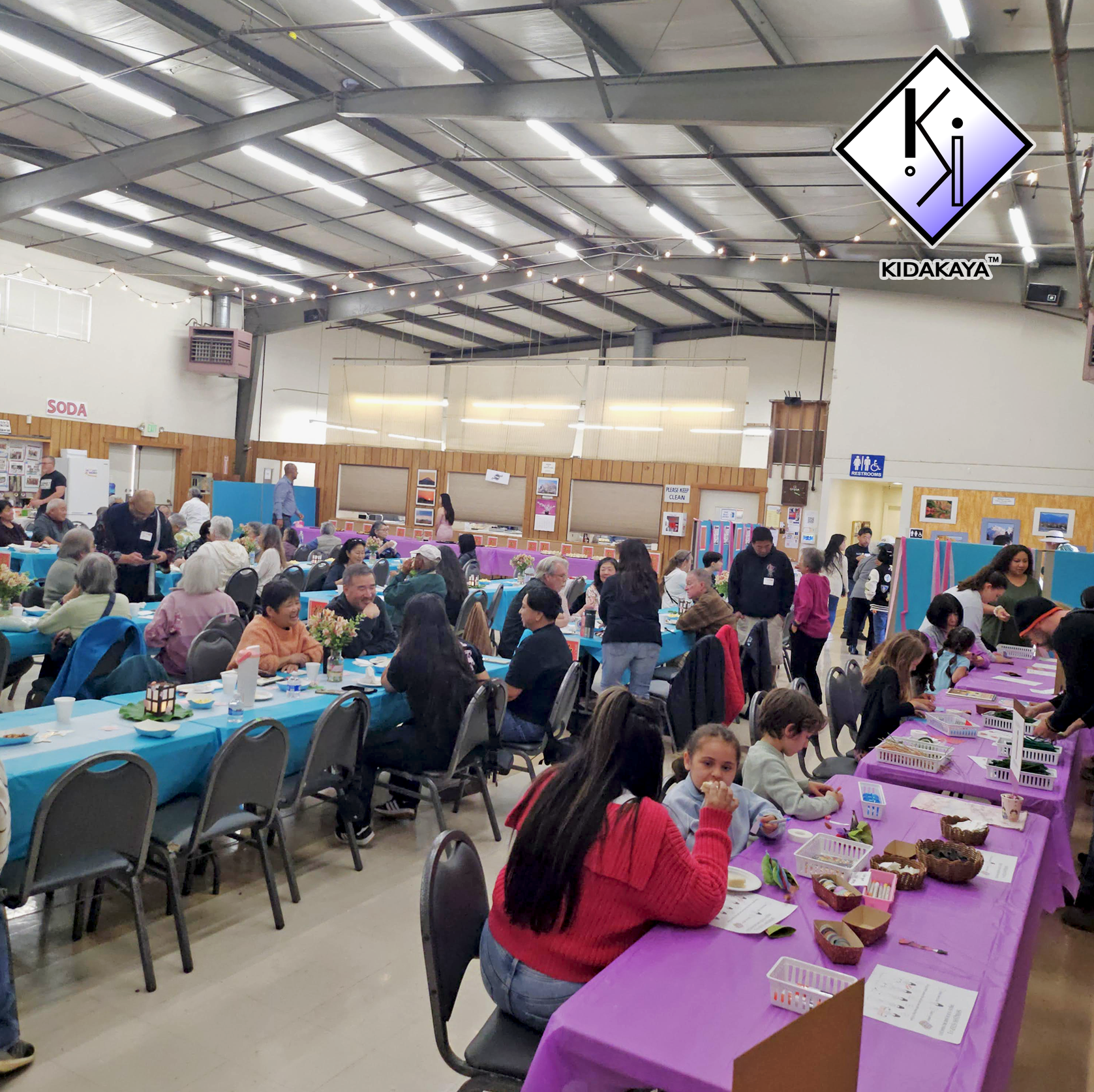 A large indoor gathering with many people seated at long tables with blue and purple tablecloths, engaging in crafts and activities, in a spacious venue with a high ceiling, string lights, and wooden accents.
