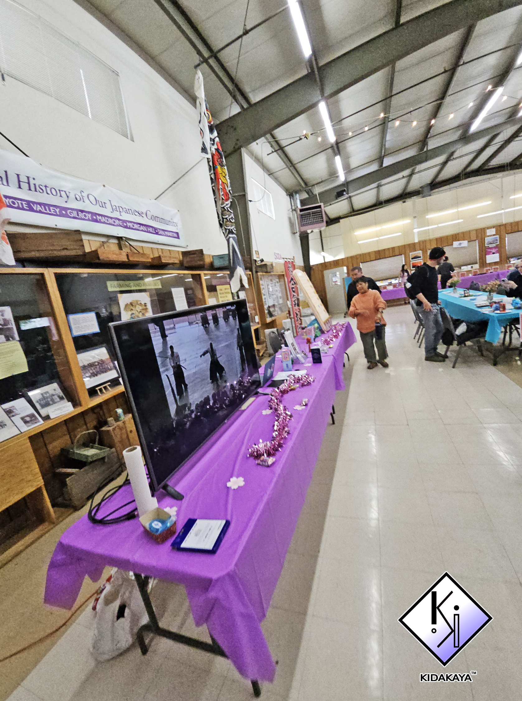A room with a high ceiling and wooden paneling on the walls, decorated for an event with multiple tables covered in purple and teal tablecloths, displaying various items and decorations; a purple table in the foreground has a flat-screen monitor, pin
