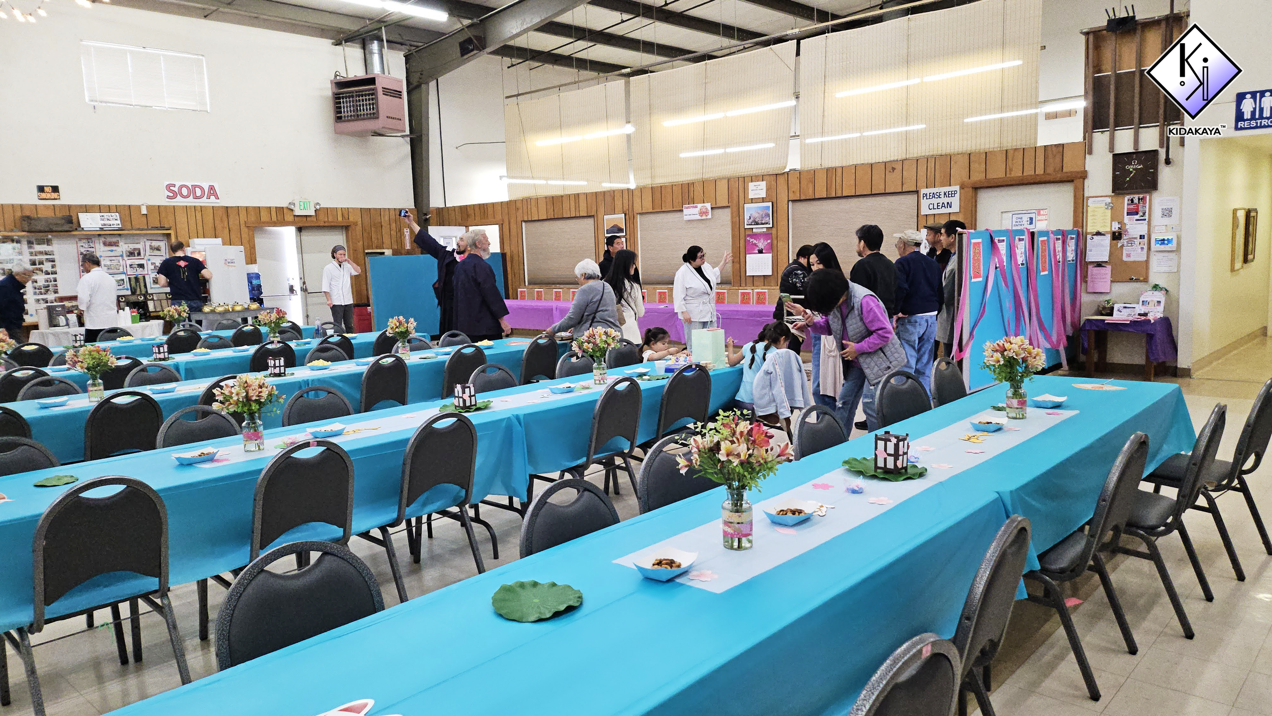Indoor community event space decorated with long tables covered in turquoise tablecloths, adorned with flower centerpieces, and set with small bowls and pink paper decorations. People are mingling and taking photos. The room has wooden walls, closed 