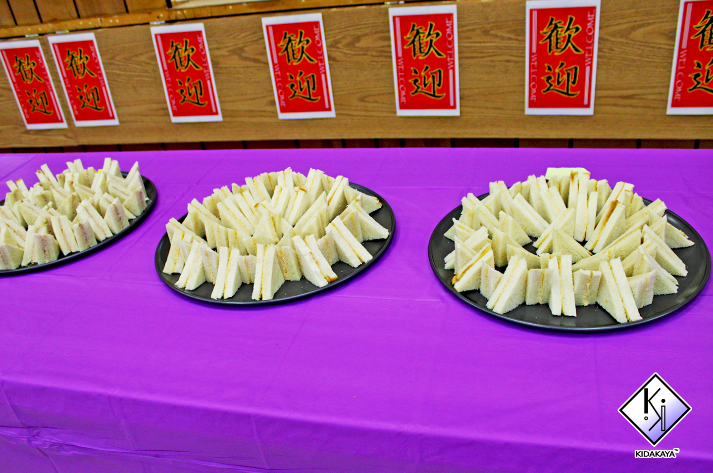 Three black platters with neatly arranged small sandwiches on a purple tablecloth, with red and yellow signs with Chinese characters and the word 'WELCOME' hanging above.