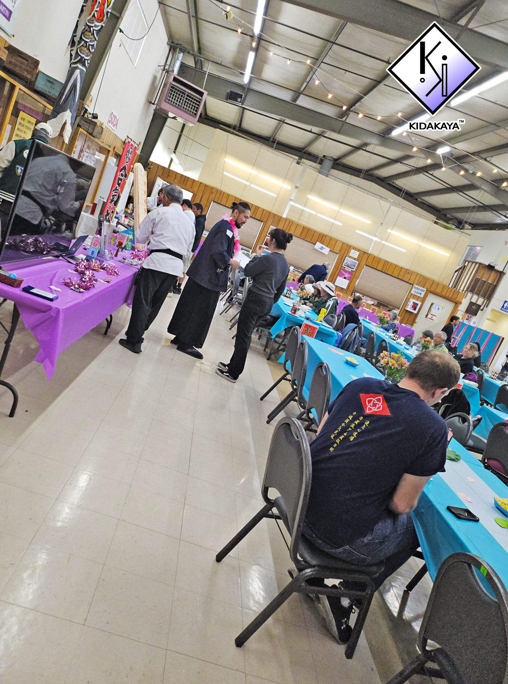 People sitting and standing in a decorated indoor space, likely for a social event or community gathering, with tables covered in blue and purple tablecloths and colorful flower arrangements.