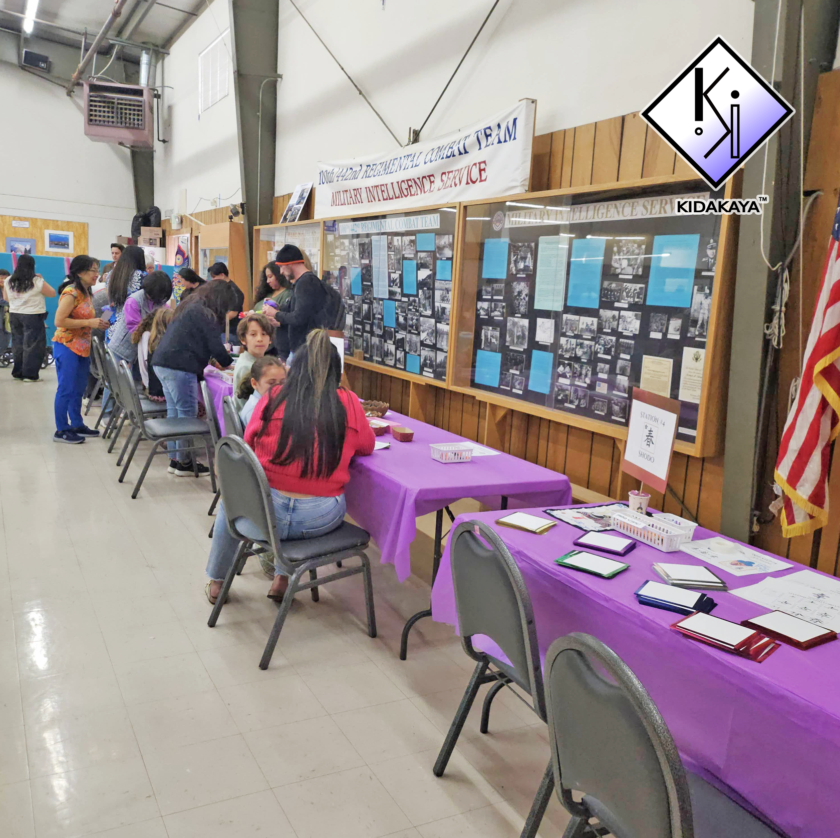 Indoor event showing a long table with purple tablecloths, chairs, and informational displays about the Military Intelligence Service. Several people are gathered around the table, some reading or talking, with display boards on the wall behind. A ba