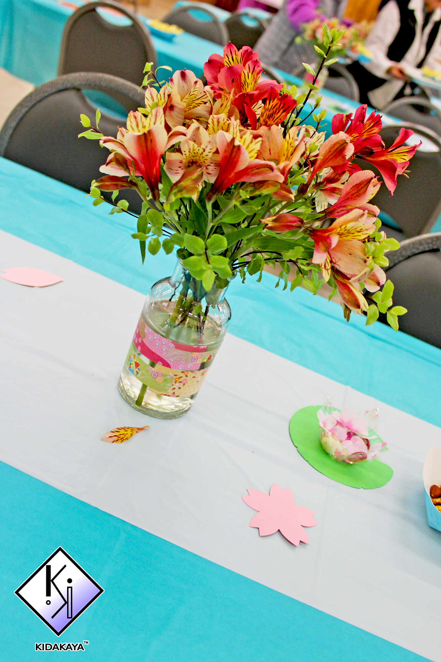 A close-up of a glass vase with a bouquet of pink, yellow, and orange flowers, including lilies, on a table decorated for an event with pink paper cutouts and a pink and green paper decoration. In the background, event chairs and tables are visible.