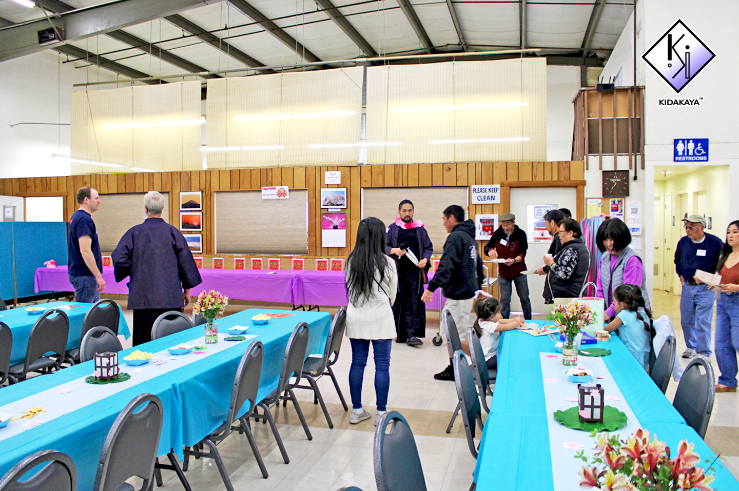 Indoor gathering with tables covered in blue tablecloths, people socializing, some children seated at a table, colorful decorations, and a wooden background with framed photos.
