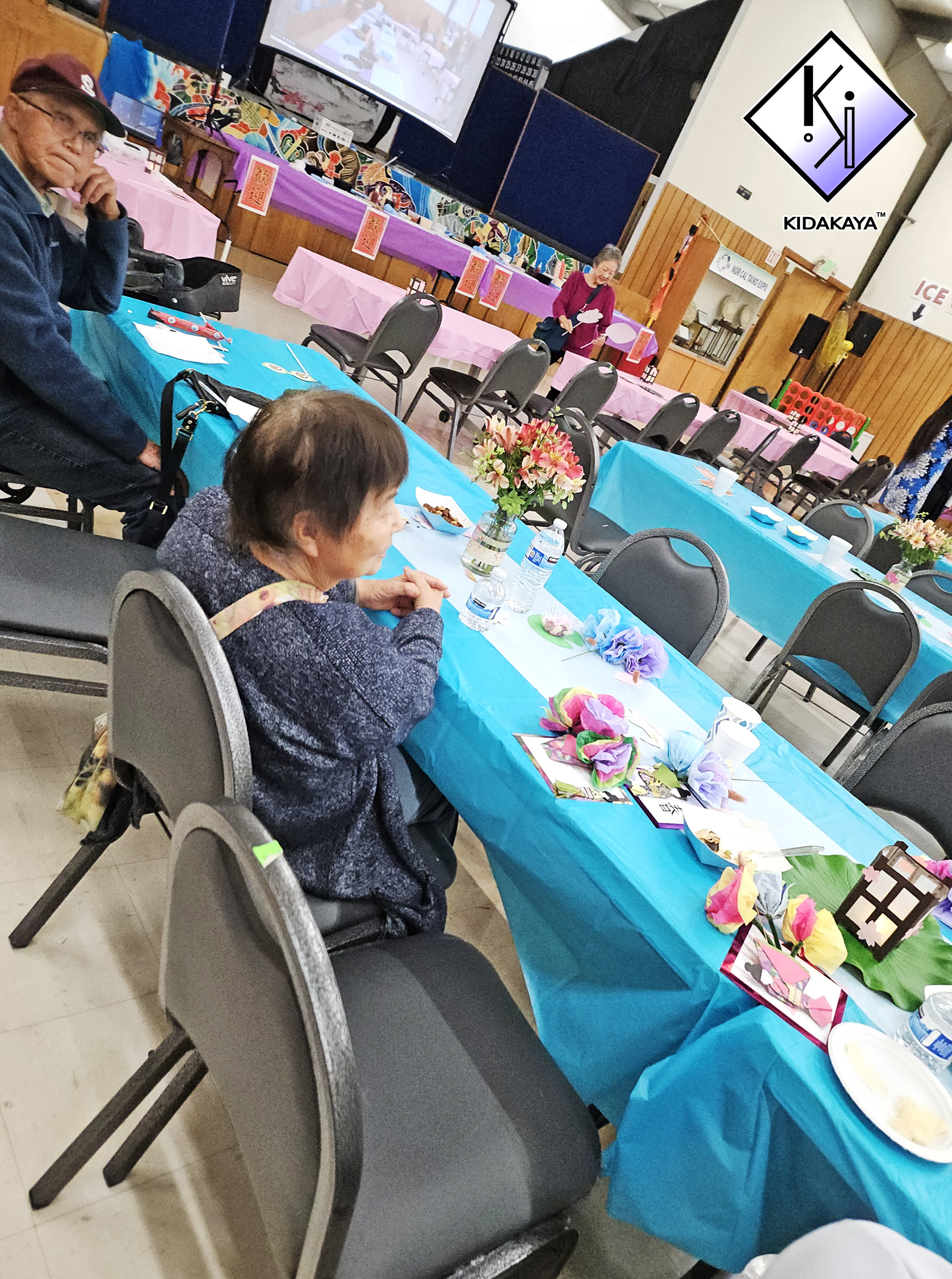 People sitting at tables decorated with pink and blue tablecloths, floral centerpieces, and paper crafts, with a woman reading at the front of a decorated room.