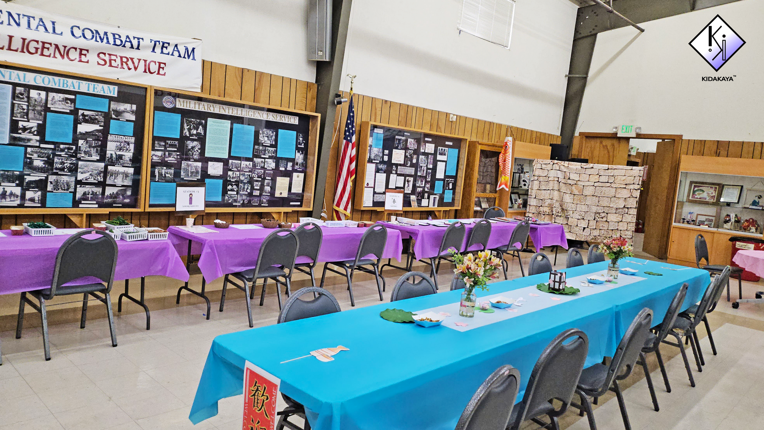 Event hall decorated with colorful tables and displays, including a purple and blue tablecloth for dining and display tables with historical photos and information about military and intelligence services. There are chairs, flowers, and banners, incl