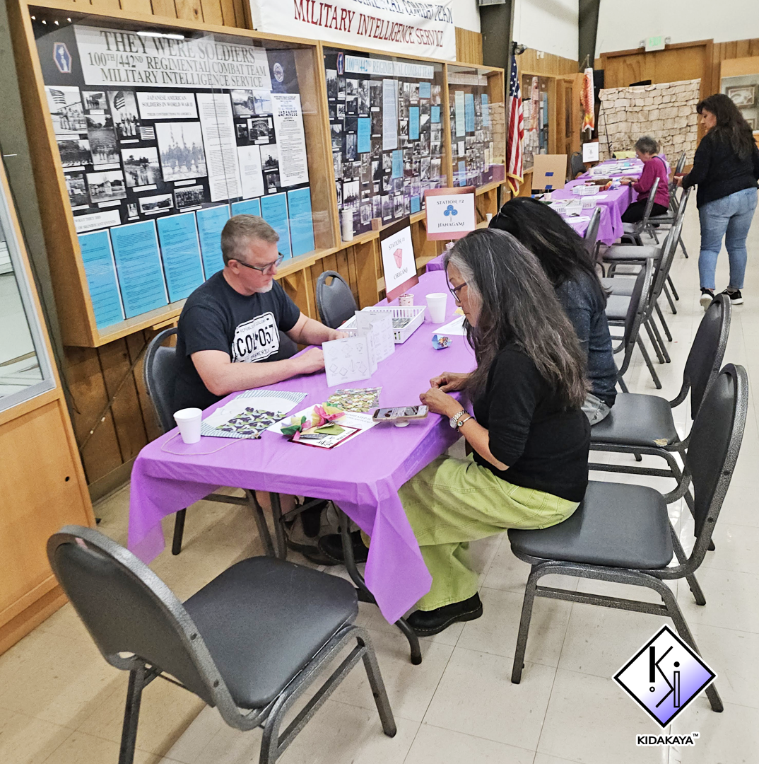 People sitting at a table with purple tablecloths, engaging in crafts or activities, in a room with wall displays about soldiers and military history, American flags, and additional tables with more activities.