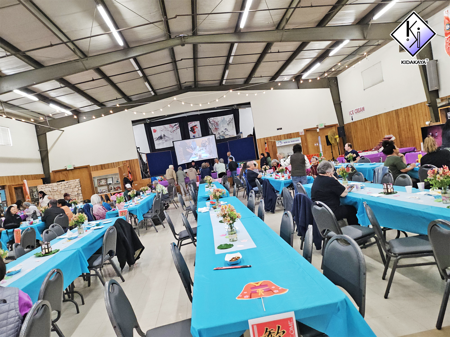 Indoor event with people seated at long tables covered with blue tablecloths, decorated with floral centerpieces, and colorful decorations, including lanterns and paper umbrellas. There is a stage with a large screen showing people seated at tables, 