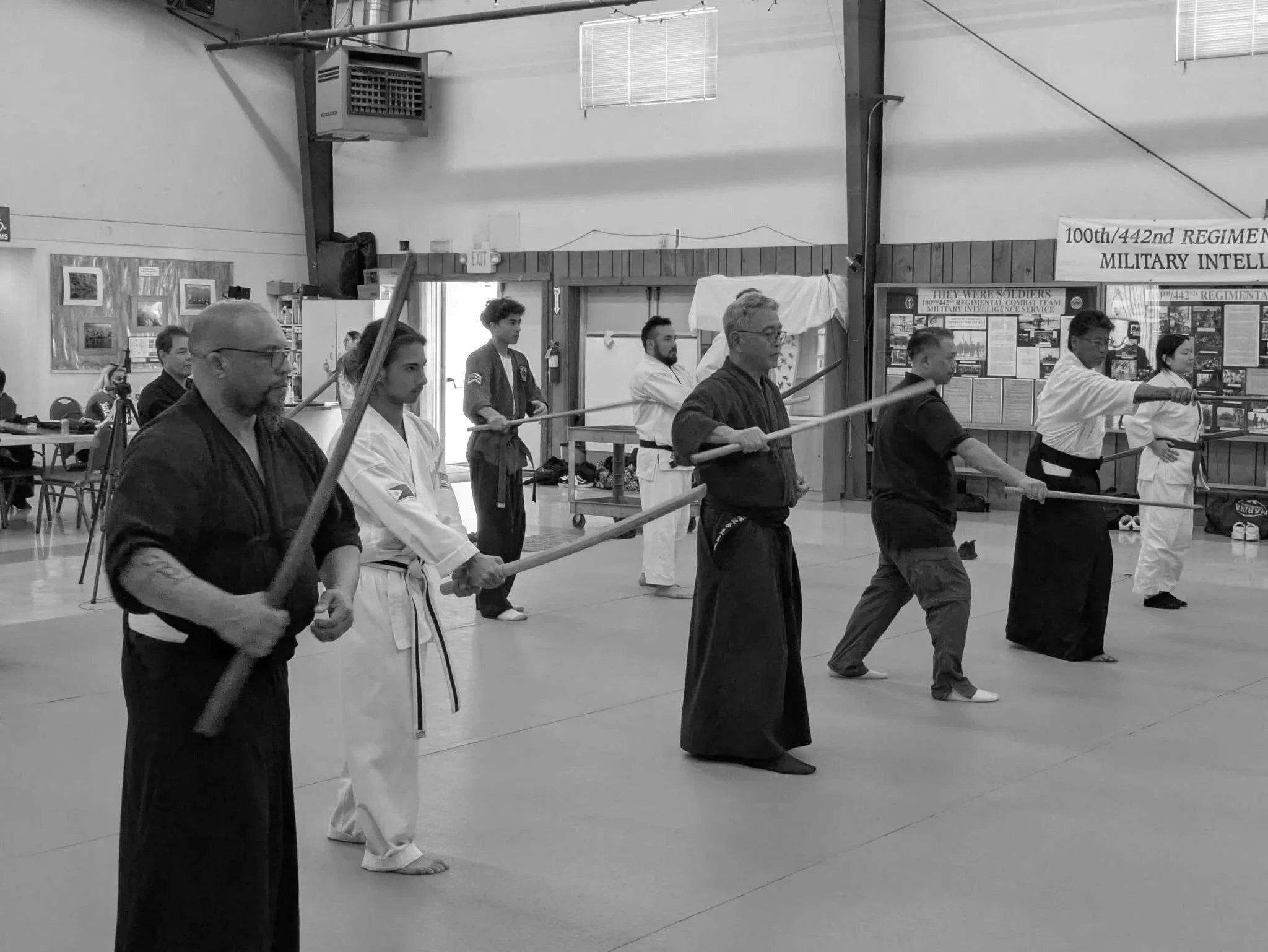 A group of martial artists, dressed in traditional uniforms called gis, stand in a line holding wooden swords as part of a martial arts class or demonstration. The room appears to be a dojo or martial arts school.