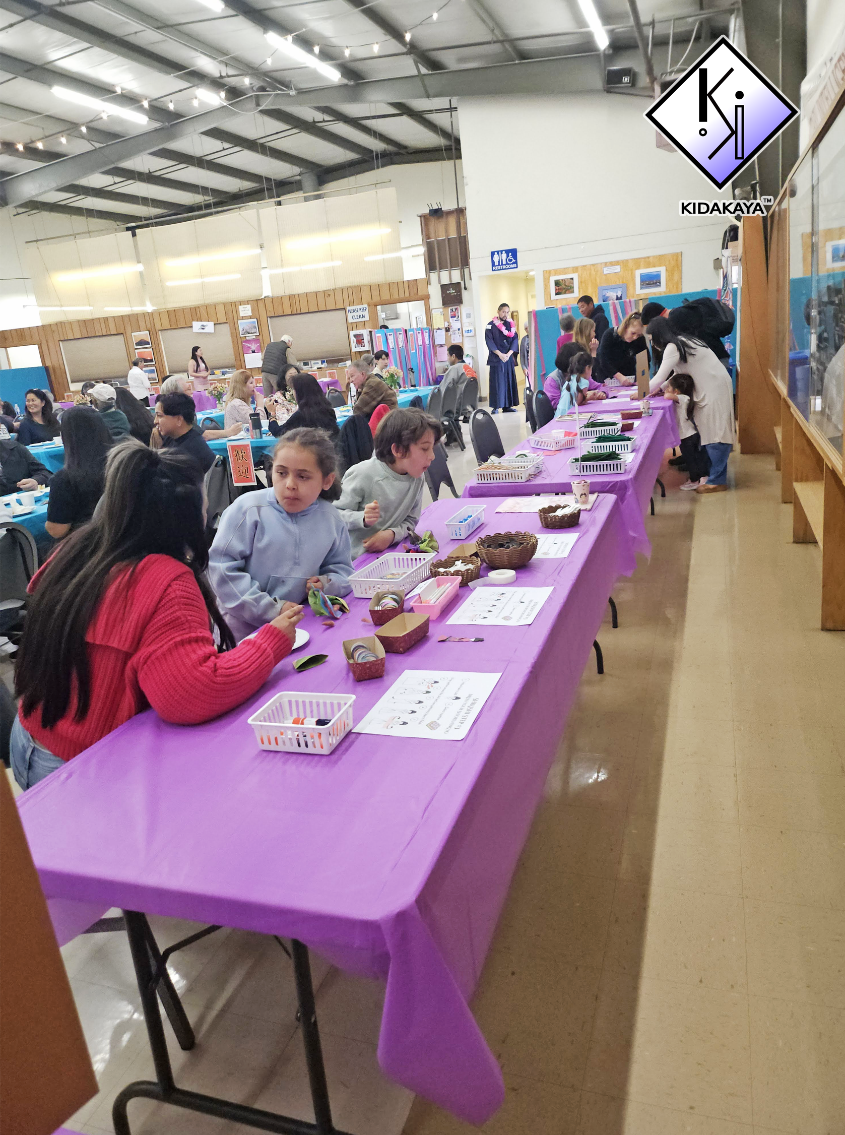 Community indoor event with various tables, some decorated with purple tablecloths, and people participating in activities. There are children and adults, some inspecting items on the tables, and a sign indicating restrooms in the background.