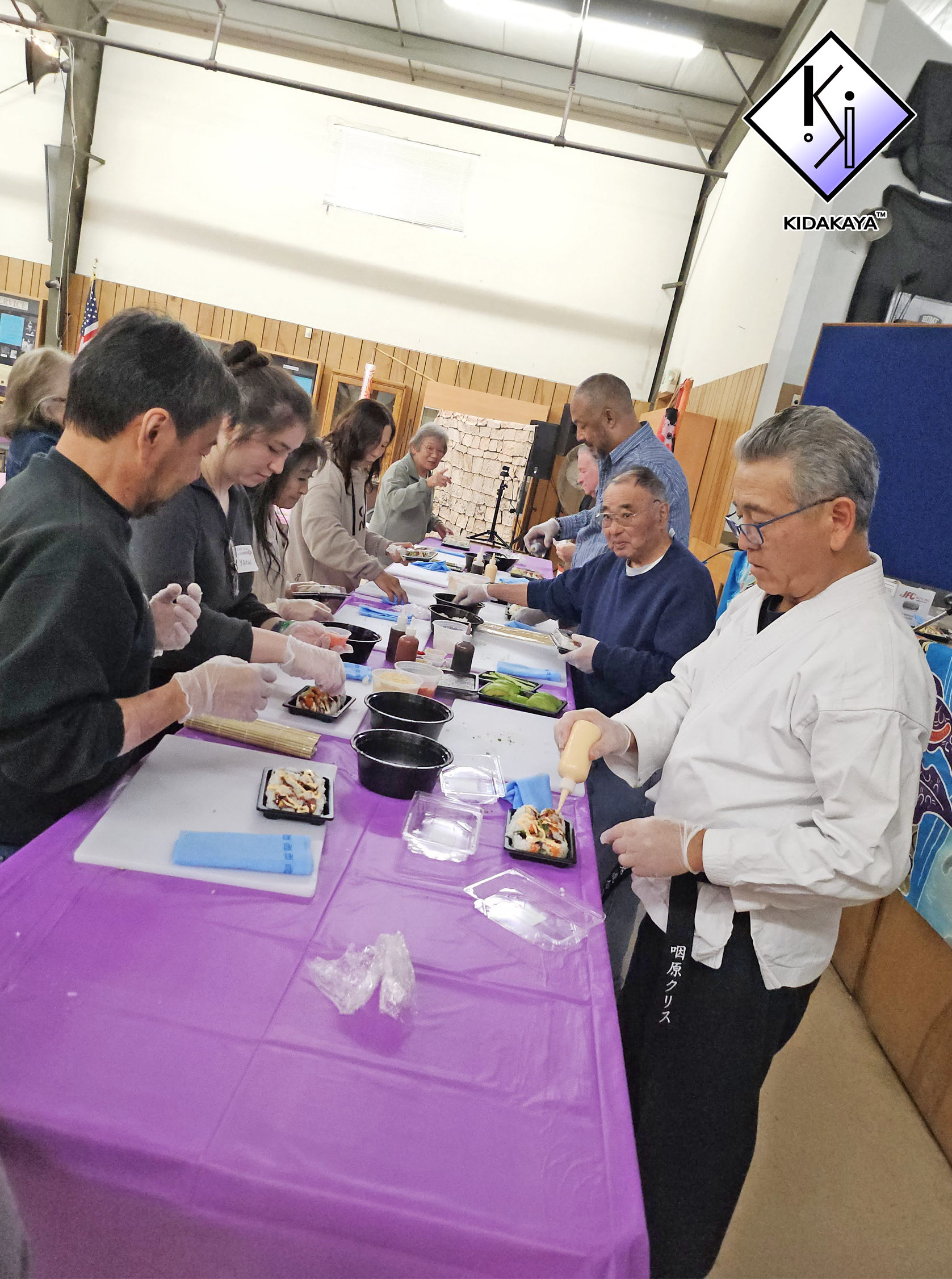 People preparing sushi at a table covered with a purple tablecloth in a room with wood-paneled walls and high ceiling.