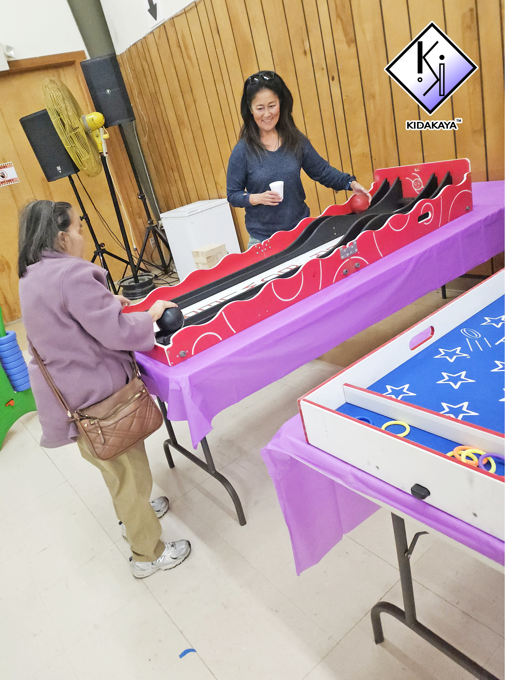 A woman in a navy long-sleeve shirt and sunglasses on her head is holding a small red ball while playing a tabletop game at a purple table. An older woman in a pink jacket with a brown handbag is watching her. The setting appears to be an indoor recr
