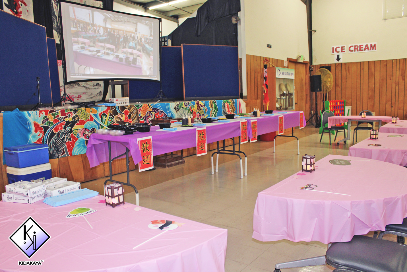 Party room decorated with pink tablecloths, lanterns, and colorful decorations, with a game setup on a stage and various tables set for an event.