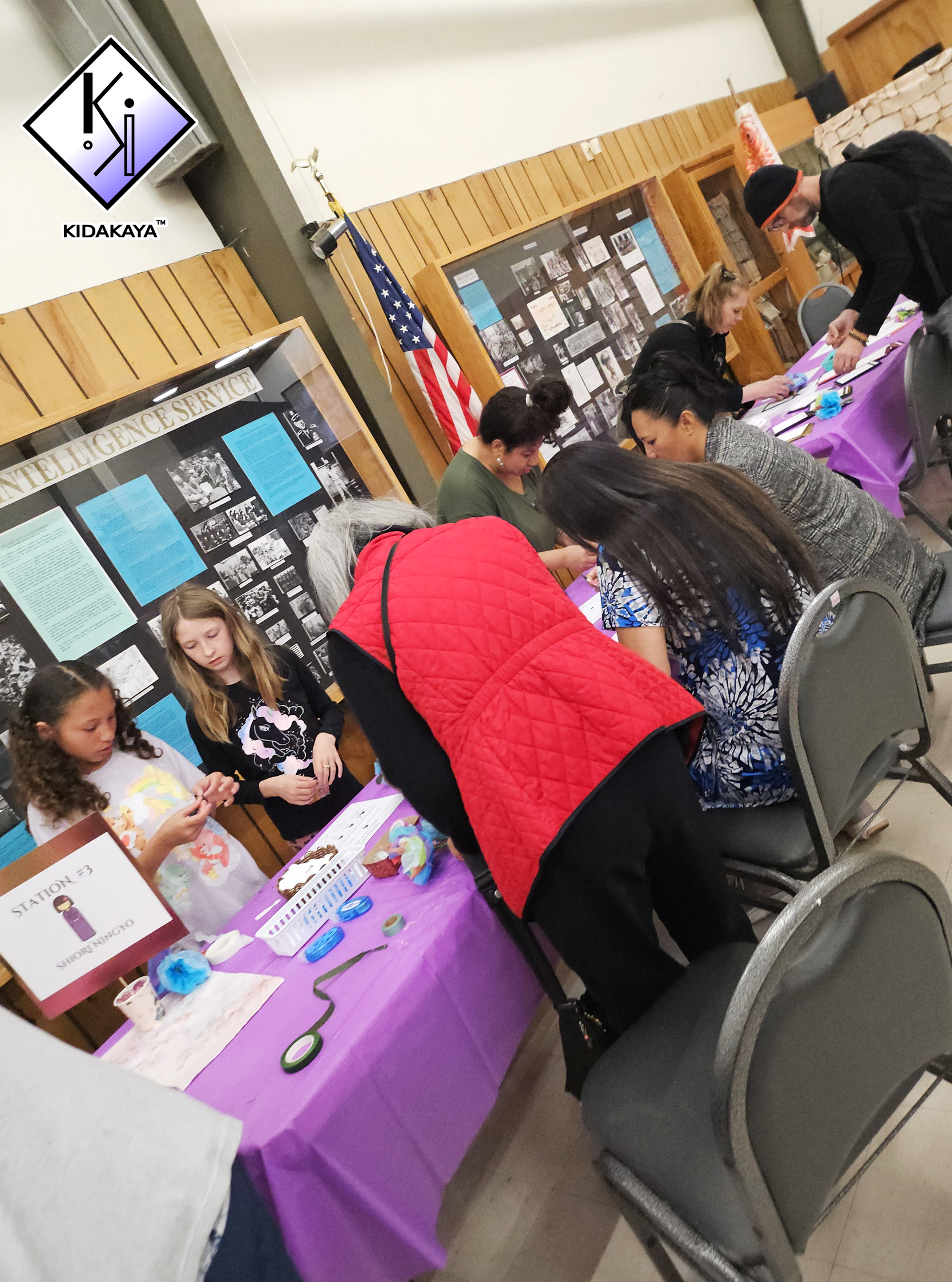 People making crafts at a table with a purple tablecloth, decorated with craft supplies, in a room with wooden paneling, display boards, and an American flag.