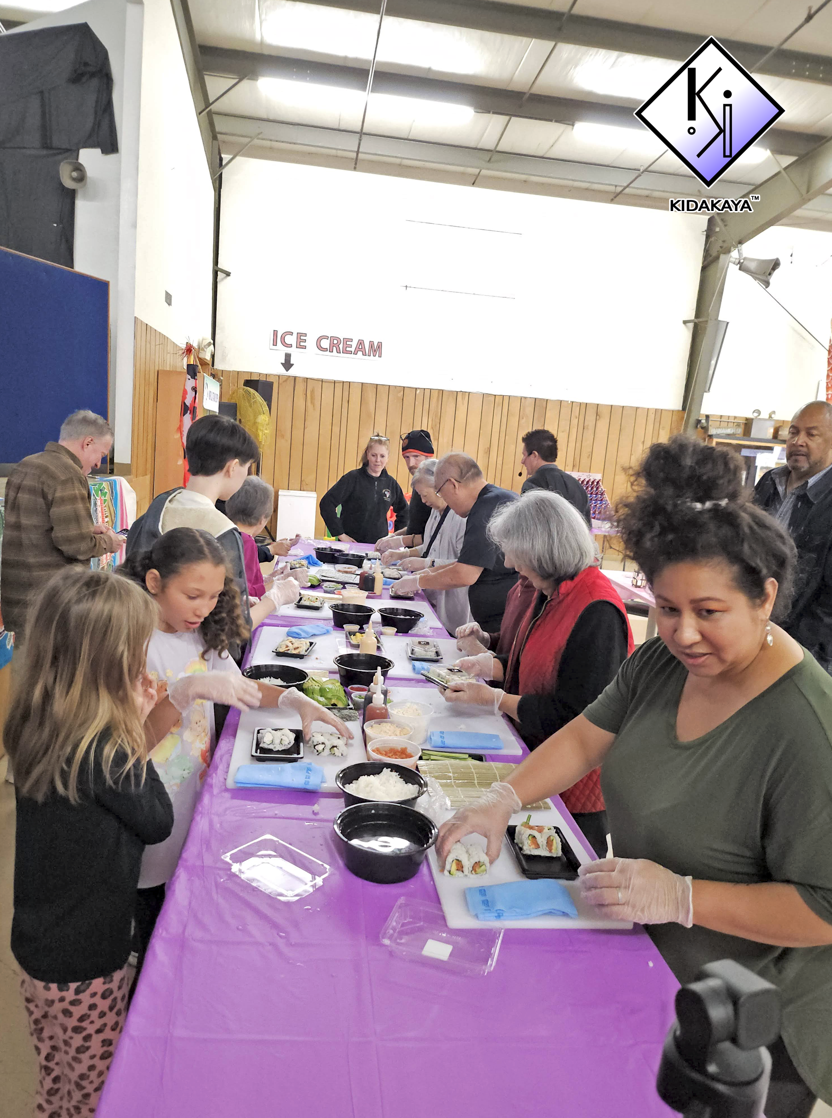 People gathered around a table making sushi in an indoor community center or event space.