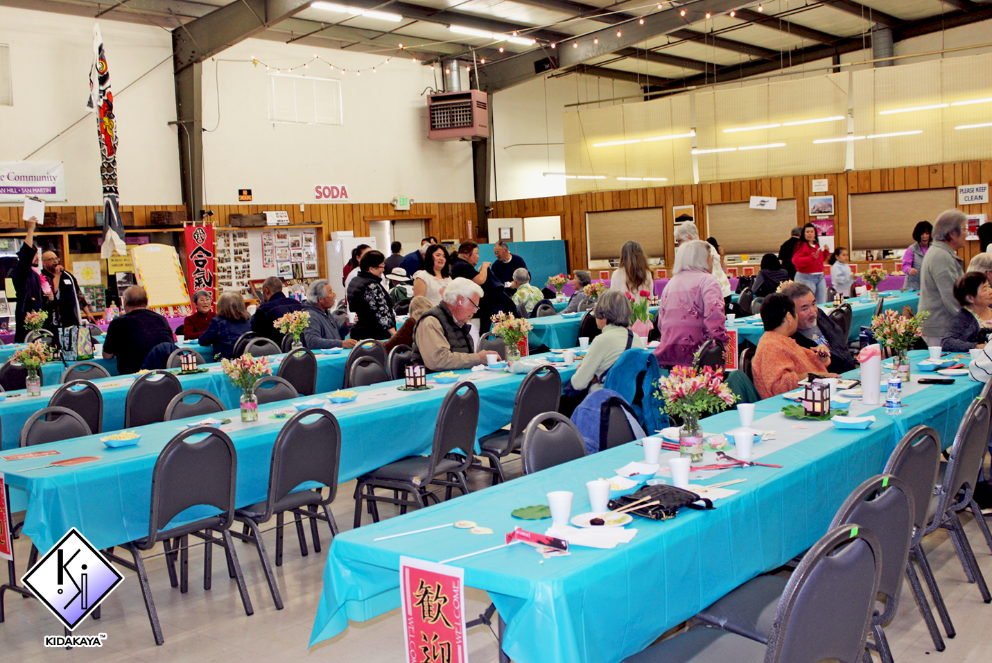 A large indoor gathering with tables covered in turquoise tablecloths, decorated with colorful flower centerpieces and set with plates, cups, and utensils, hosting diverse people socializing and dining.