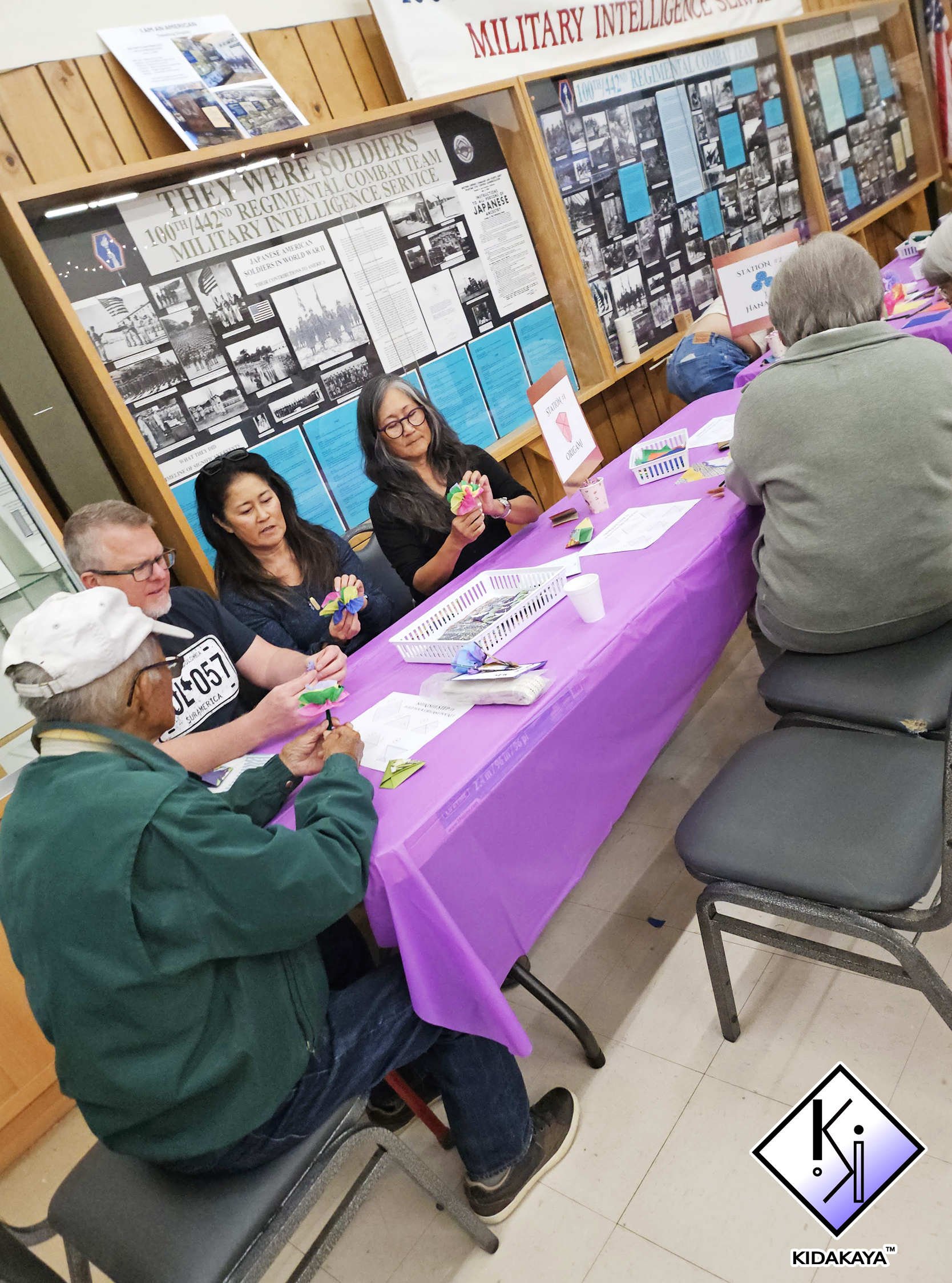 People participating in a craft activity at a community event, seated at a purple table with craft supplies, with informational posters and display boards about military history on the wall behind them.