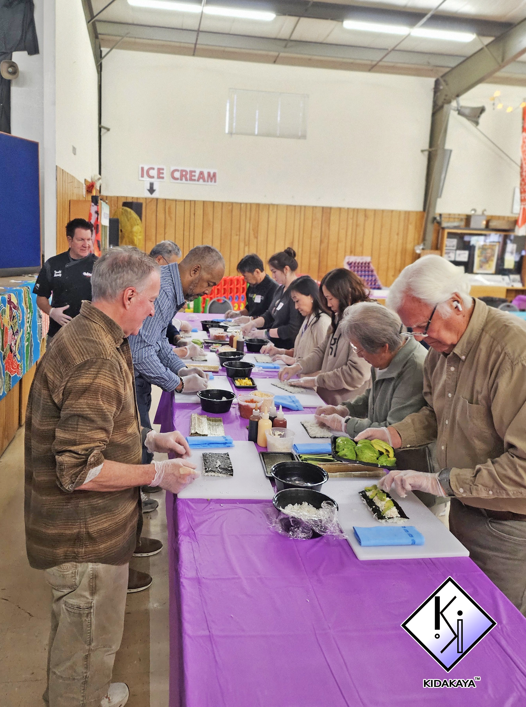 People participating in a sushi-making class, standing along a long table covered with a purple tablecloth, preparing and rolling sushi.