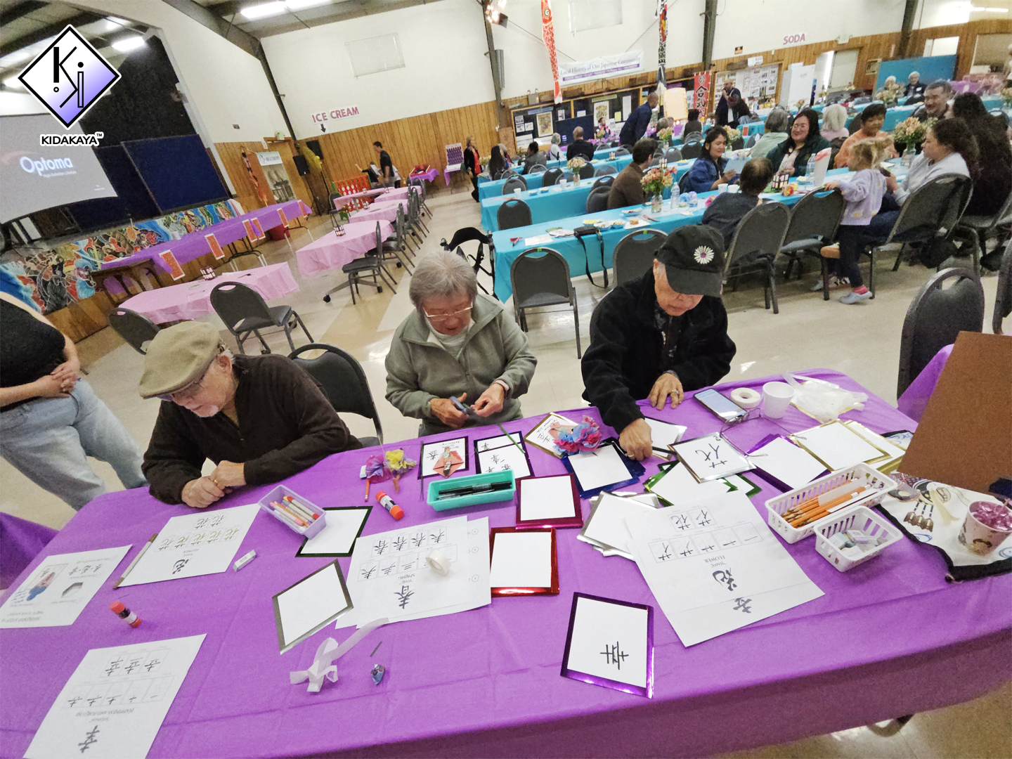 People sitting at a table with papers, markers, and picture frames, participating in a calligraphy or writing activity in a decorated community hall with tables, chairs, and other groups in the background.