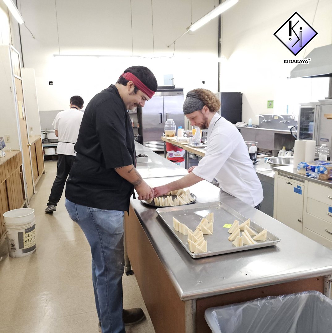 Two chefs, one wearing a black shirt and red headband, and the other in a white chef's coat and gray headband, are preparing sandwiches in a commercial kitchen. There are trays of sliced bread on the counter, and another chef is working in the backgr