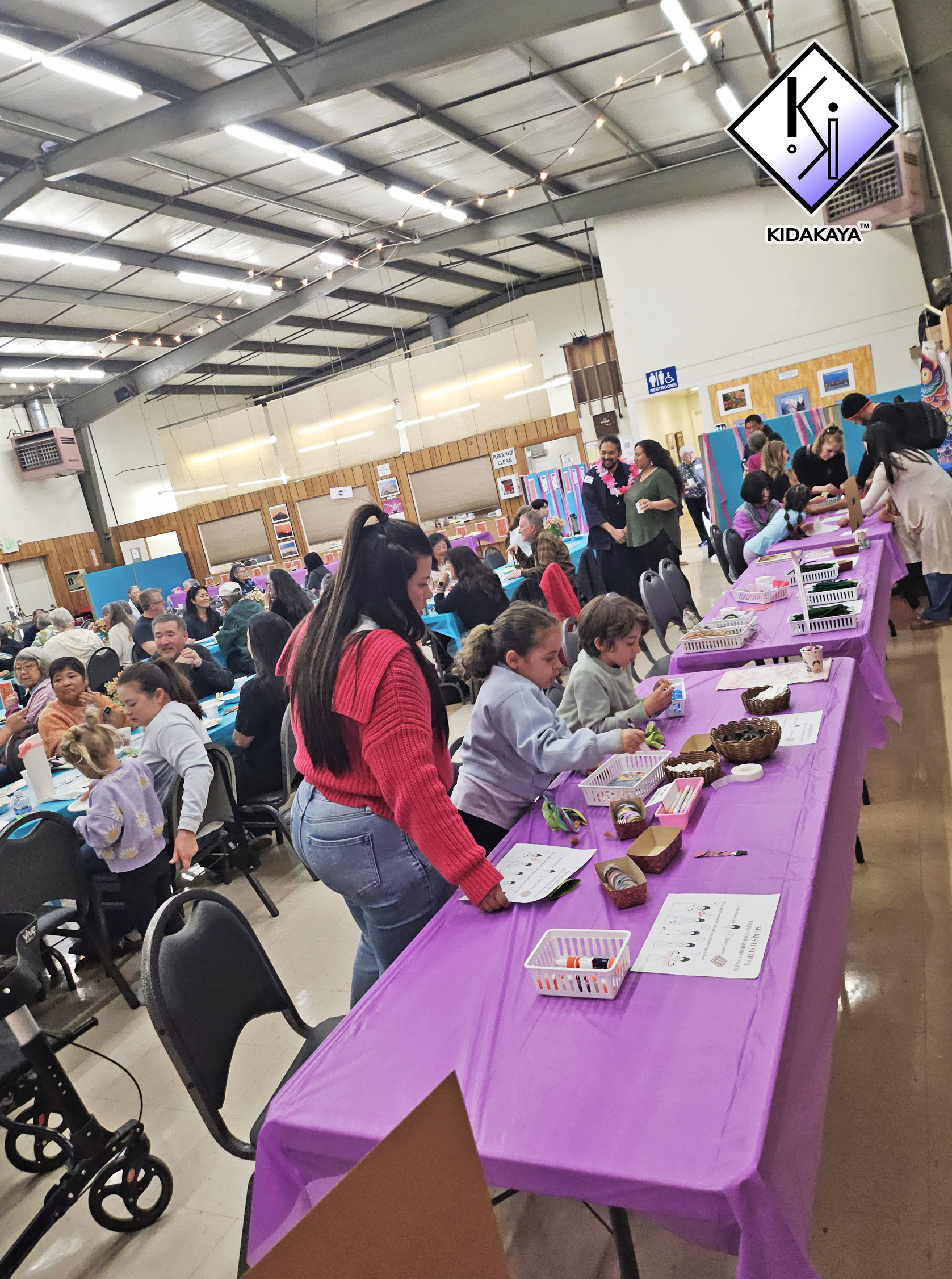 A community event with people gathered around decorated tables, participating in arts and crafts, in a spacious indoor venue with high ceilings and string lights.