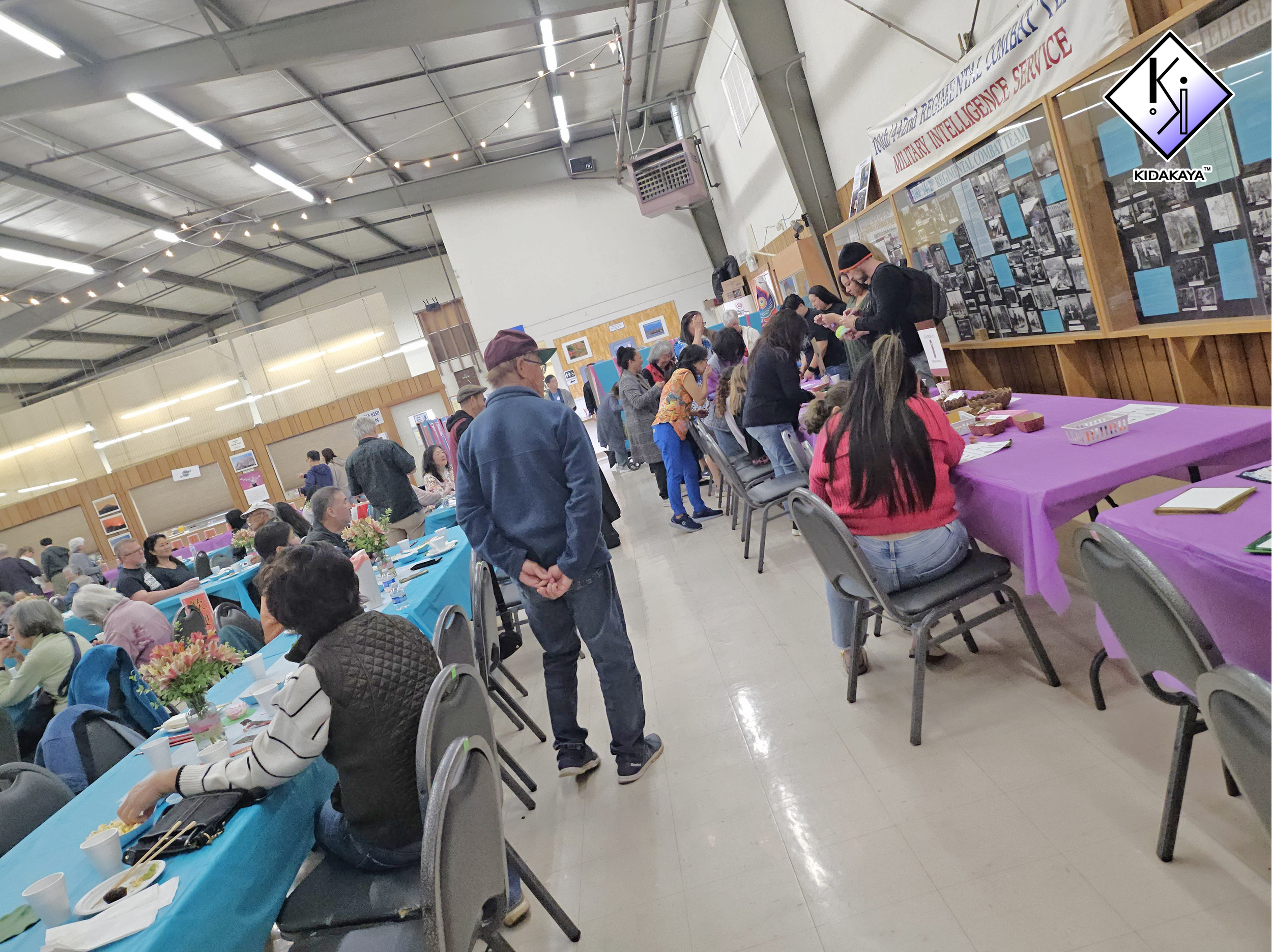 People gathered at tables with purple and blue tablecloths inside a hall, participating in an event or celebration.