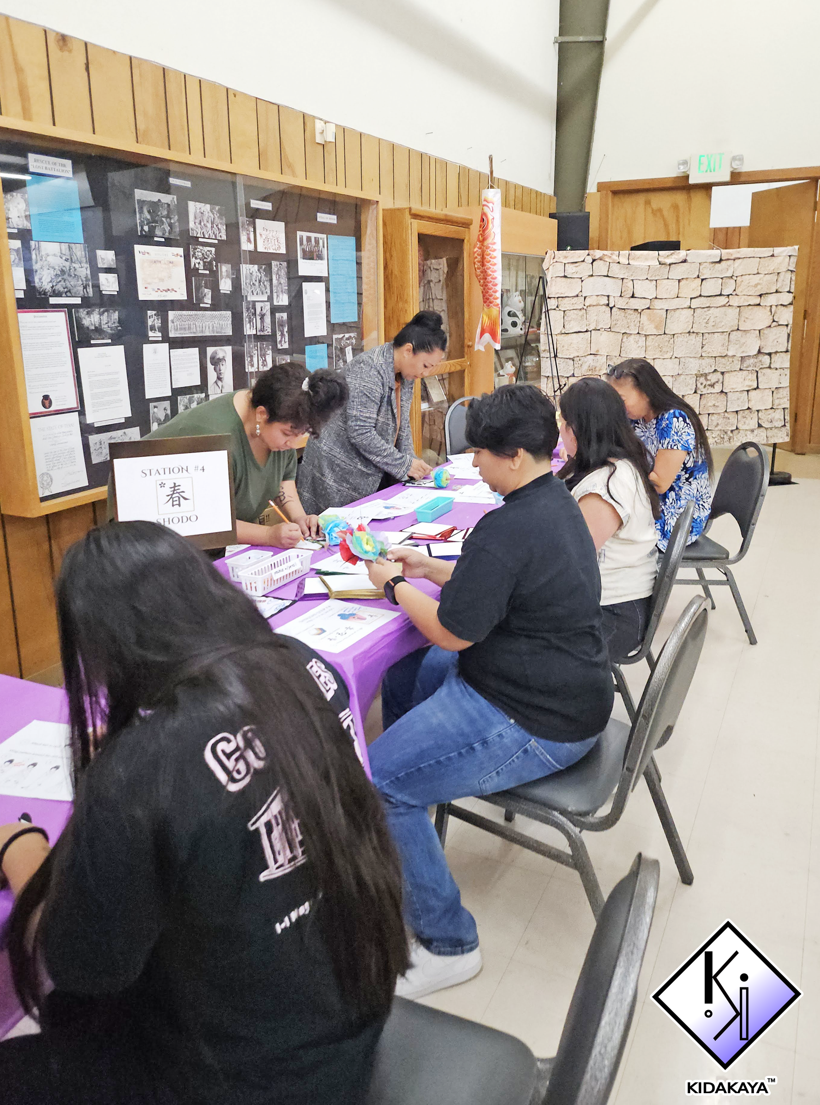 Group of people sitting and standing around a table in a room with wooden walls, working on arts and crafts projects with paper and colorful materials.