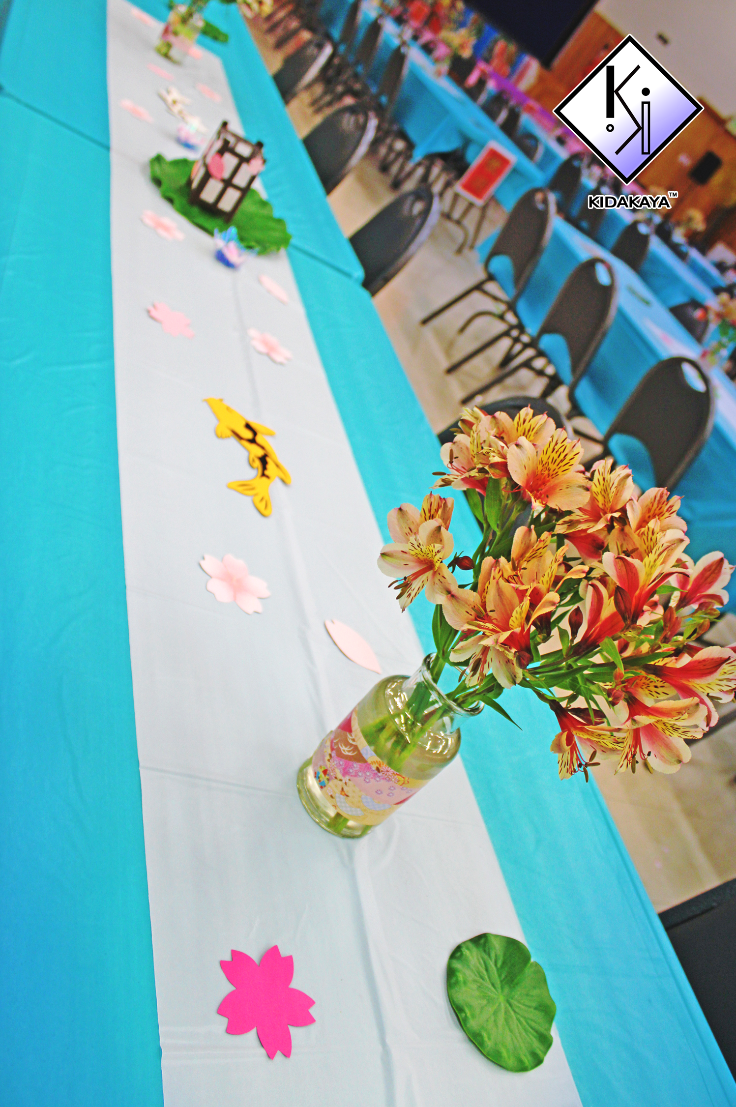 Decorated table with pink and yellow lilies in a glass vase, artificial green leaves, pink paper cutouts of flowers, a small black and white gift box, and gray chairs arranged around the table in a decorated event space.