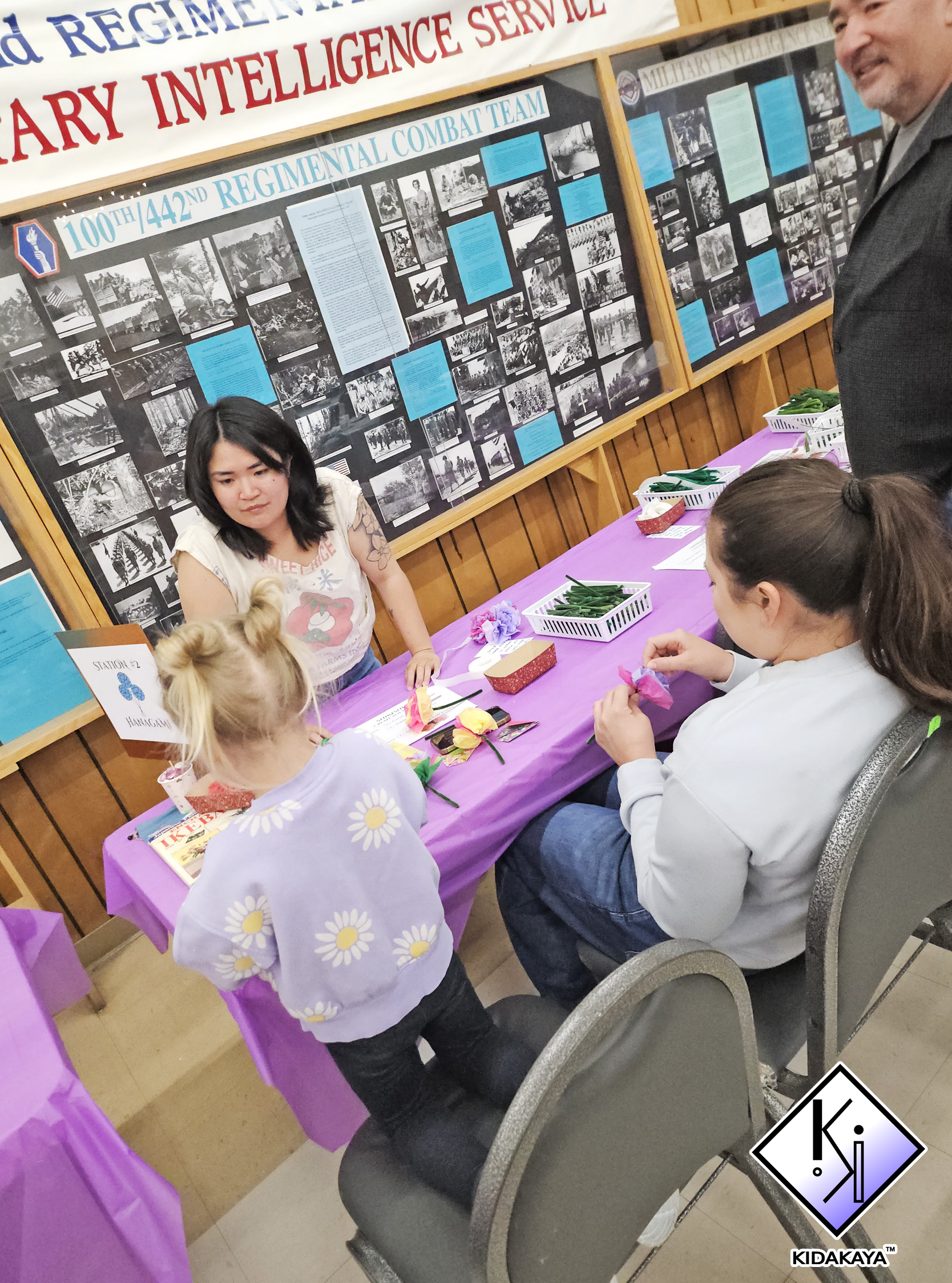 A classroom or community event setting with children and adults around a table covered with a purple tablecloth. The table has small baskets with green leaves, booklets, and flowers. There are black and white historical photographs and text on a larg