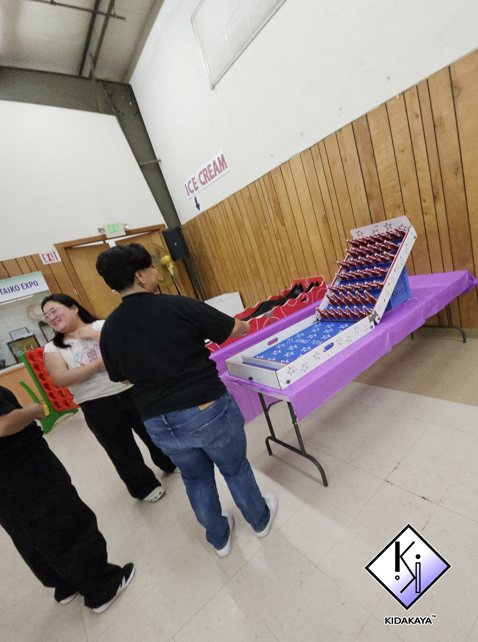 People standing in line at a table with donuts and beverages in a community hall with wooden wall paneling and signs for ice cream and exits.