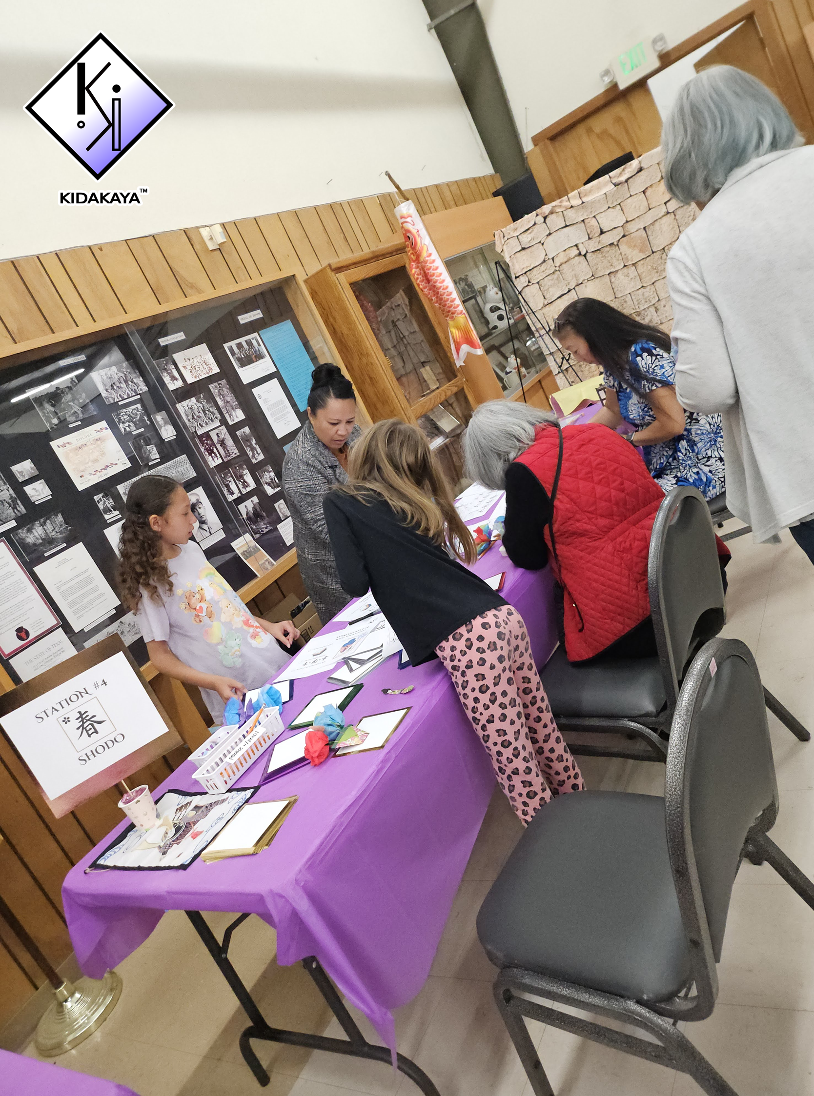 A group of people gathered around a table with a purple tablecloth, engaging in craft activities. There are informational posters and crafts on display, with a sign indicating 'Station #4' and containing Japanese characters for 'spring' and 'Shodo.' 
