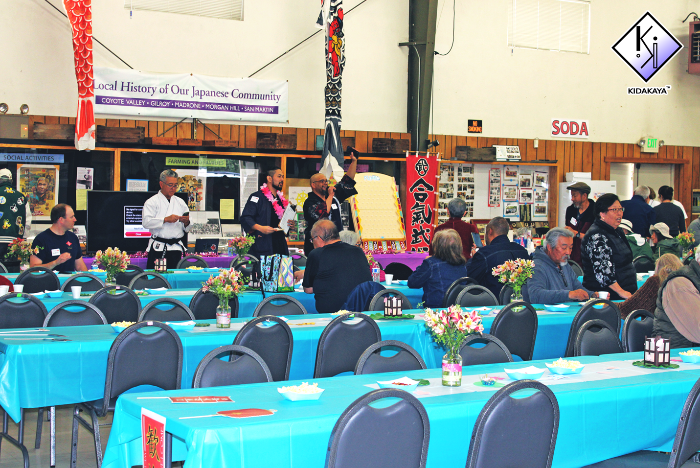 People gathered in a community hall for a cultural event, with tables covered in blue tablecloths, decorated with flowers, and a stage with speakers and posters in the background.