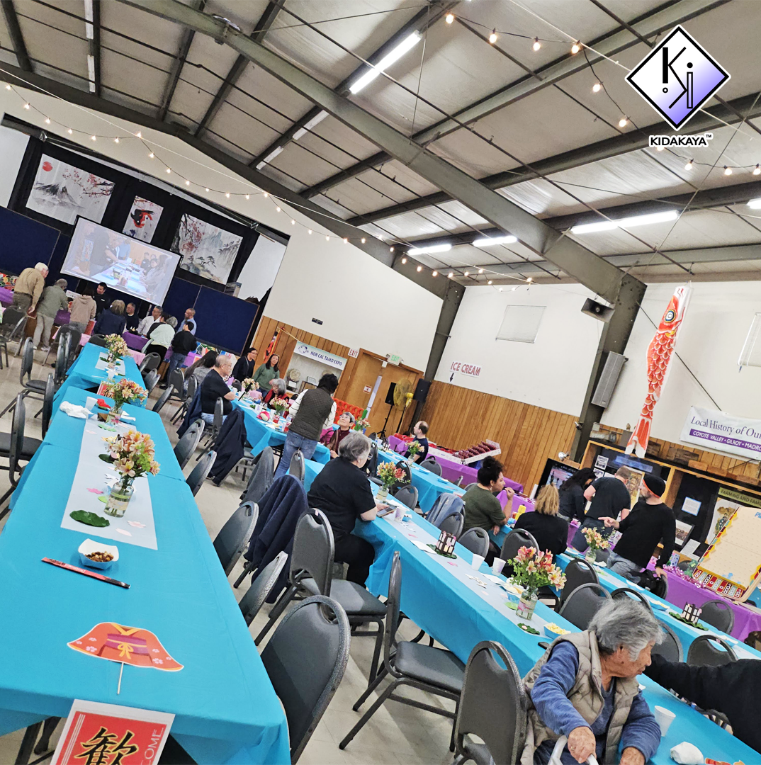 Inside of a community hall decorated for a celebration with blue tables, floral centerpieces, and people seated and standing, attending an event with a stage, banners, and a large screen.