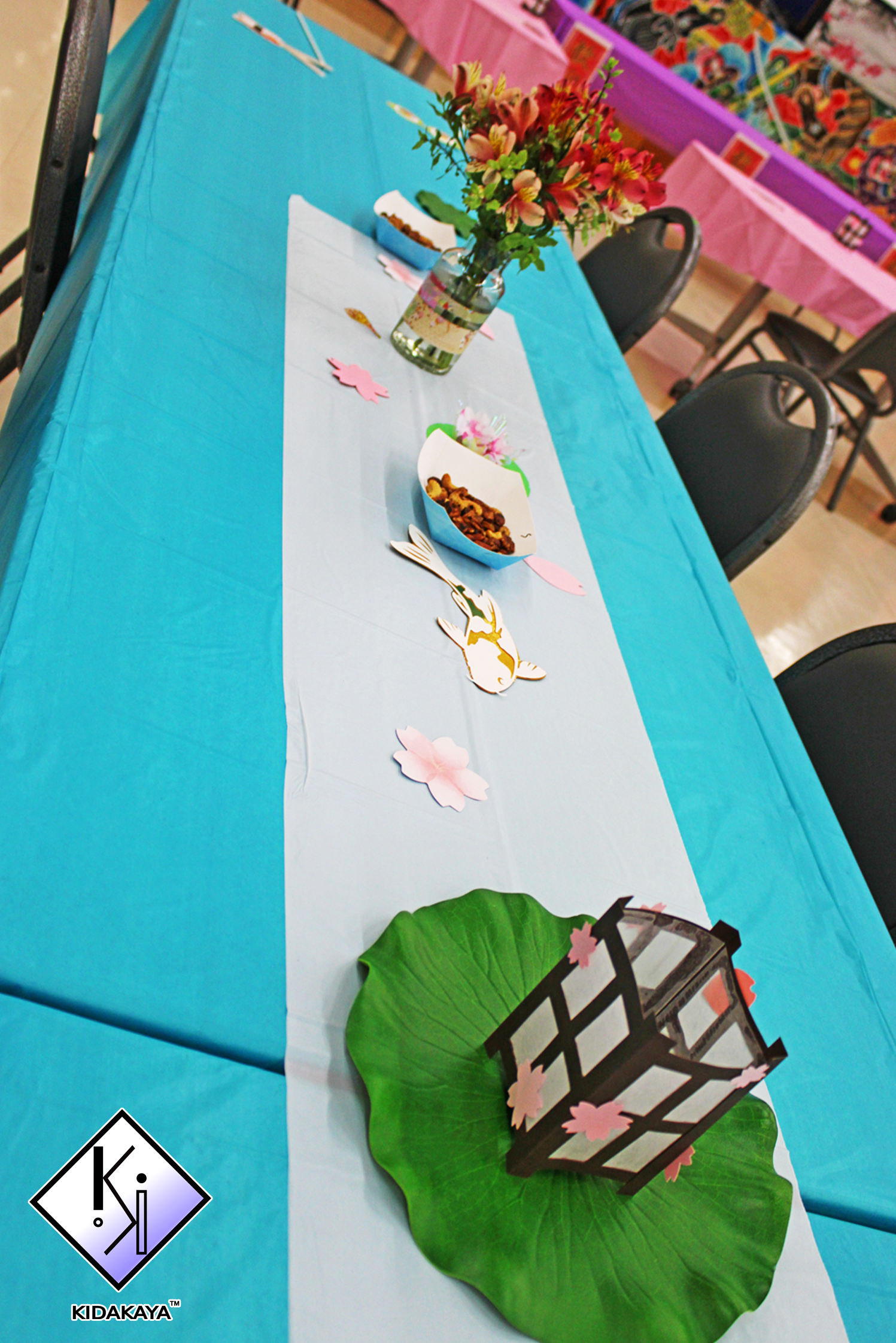 A decorated table with a blue tablecloth, a vase of pink and orange flowers, paper cutouts of pink flowers and a paper lantern with cherry blossom decorations, and a large green leaf. In the background, tables with pink tablecloths and colorful artwo
