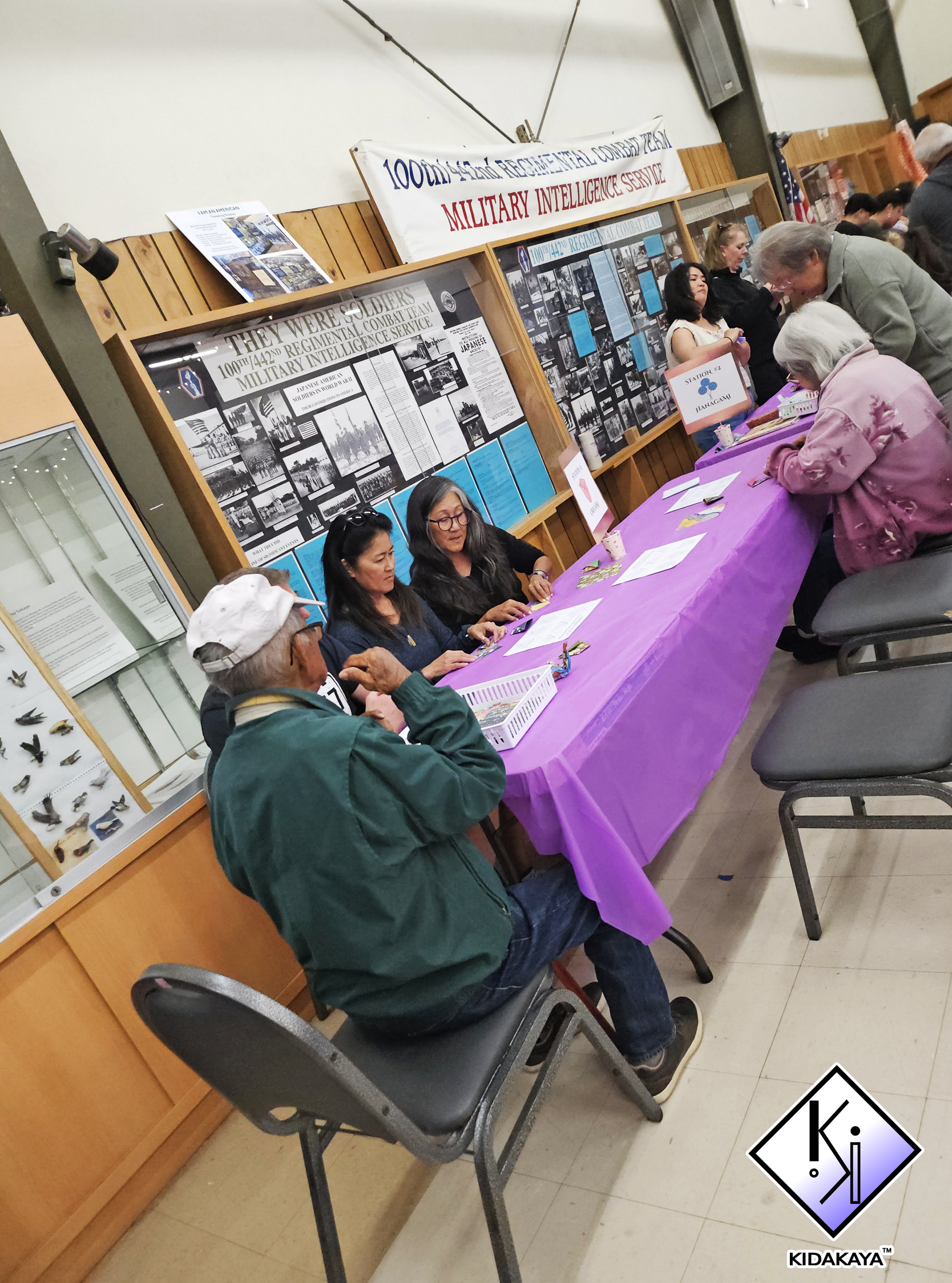 People at an informational display and game booth at an event celebrating the 100th 442nd Regimental Combat Team, with a banner reading '100th/442nd Regimental Combat Team Military Intelligence Service'.