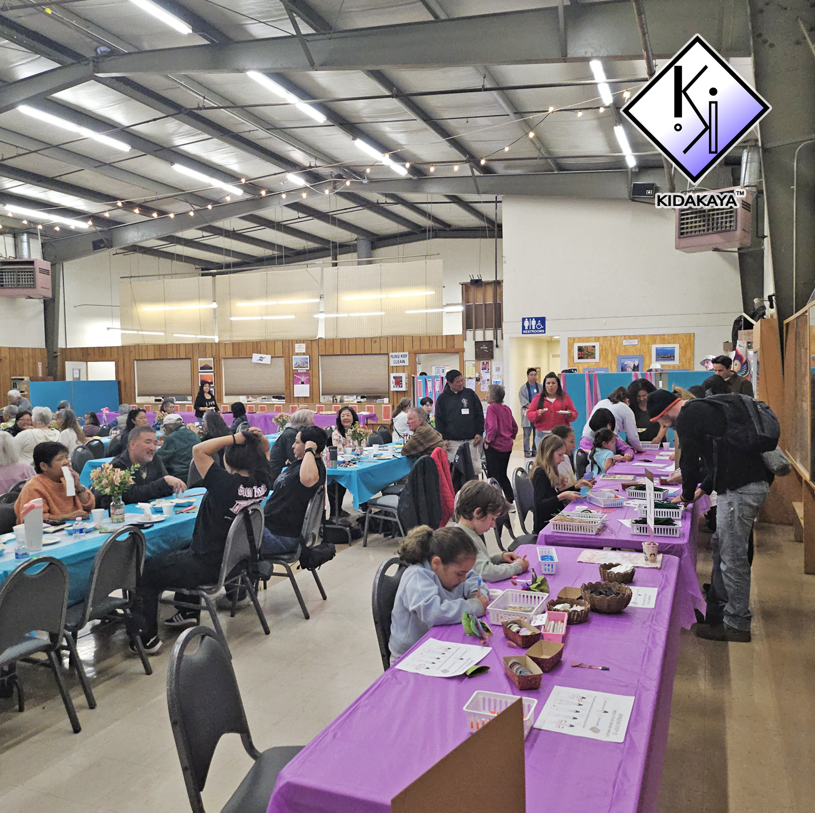 A crowded indoor event with people seated at tables decorated with blue and purple tablecloths, people standing and interacting, and craft supplies on purple tables. There are restroom signs and artwork on the walls, with a high ceiling and industria