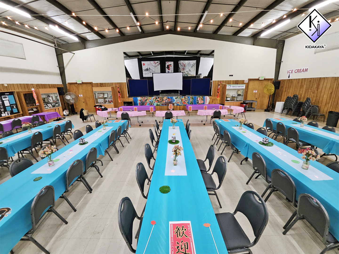 An event hall decorated with blue and pink tablecloths, small floral centerpieces, and displayed banners, with a stage at the front and chairs stacked against the walls.