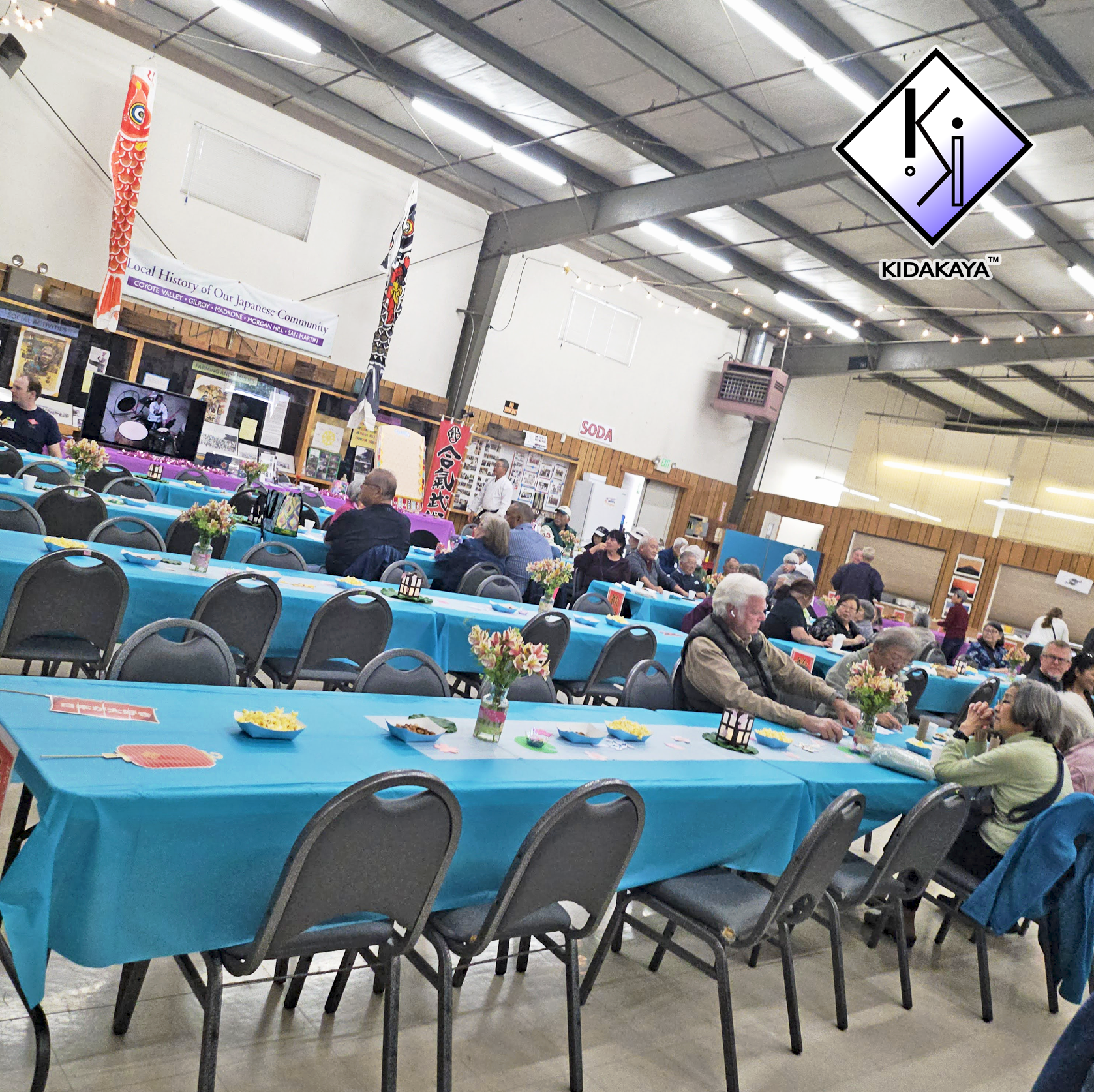 A community event in a large hall with blue tablecloth-covered tables decorated with small floral centerpieces. People are seated, socializing and enjoying snacks, with a few standing and walking around. There are banners and posters about Japanese c