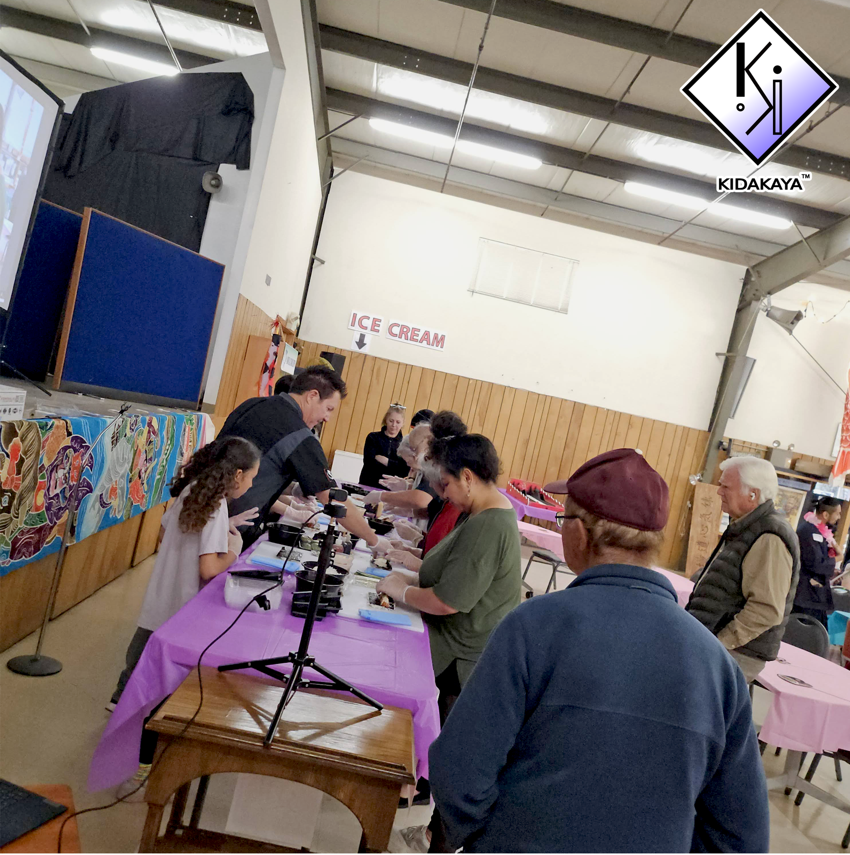 People gathered at a table with microscopes, engaging in a hands-on activity, inside a large room with a sign pointing to ice cream. There are multiple individuals, including children and adults, participating in the event.