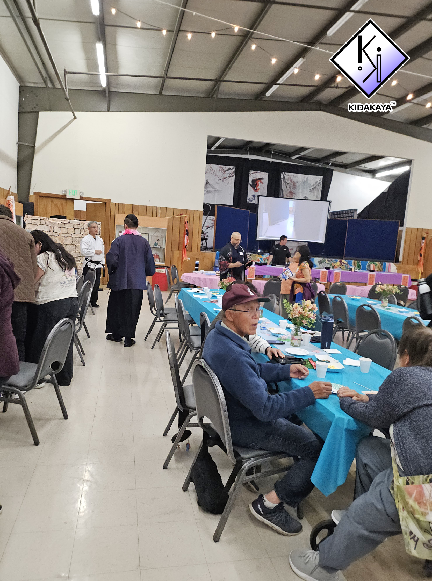 People sitting at tables with blue tablecloths at a community event or celebration, with decorations and a large screen in the background.