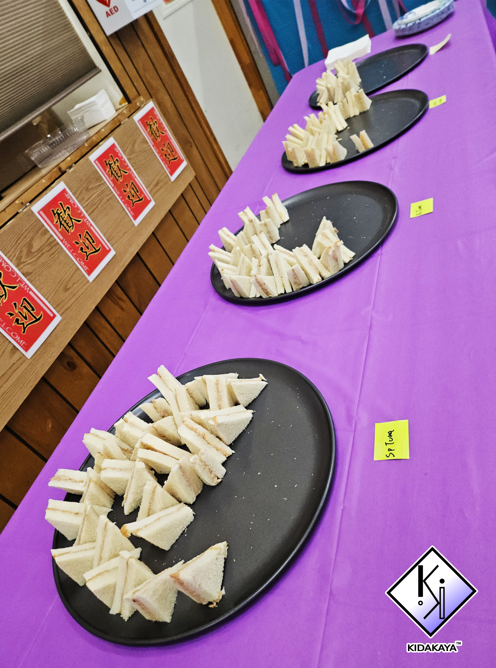 Three black trays with neatly cut sandwich triangles on a purple tablecloth, in front of a wooden wall with Chinese character posters and an emergency AED sign.