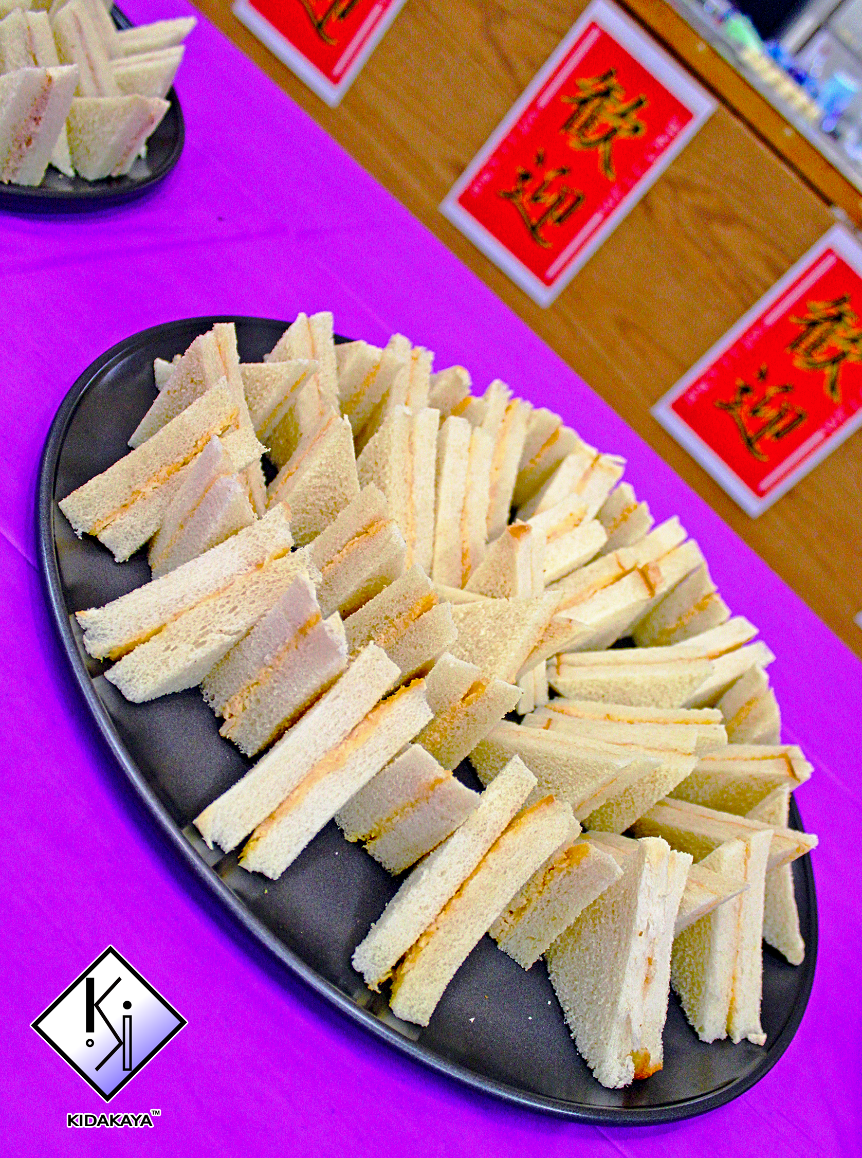 Plate of small finger sandwiches with white bread and light filling, arranged in a circle on a black platter with purple tablecloth, and Chinese character signs in the background.