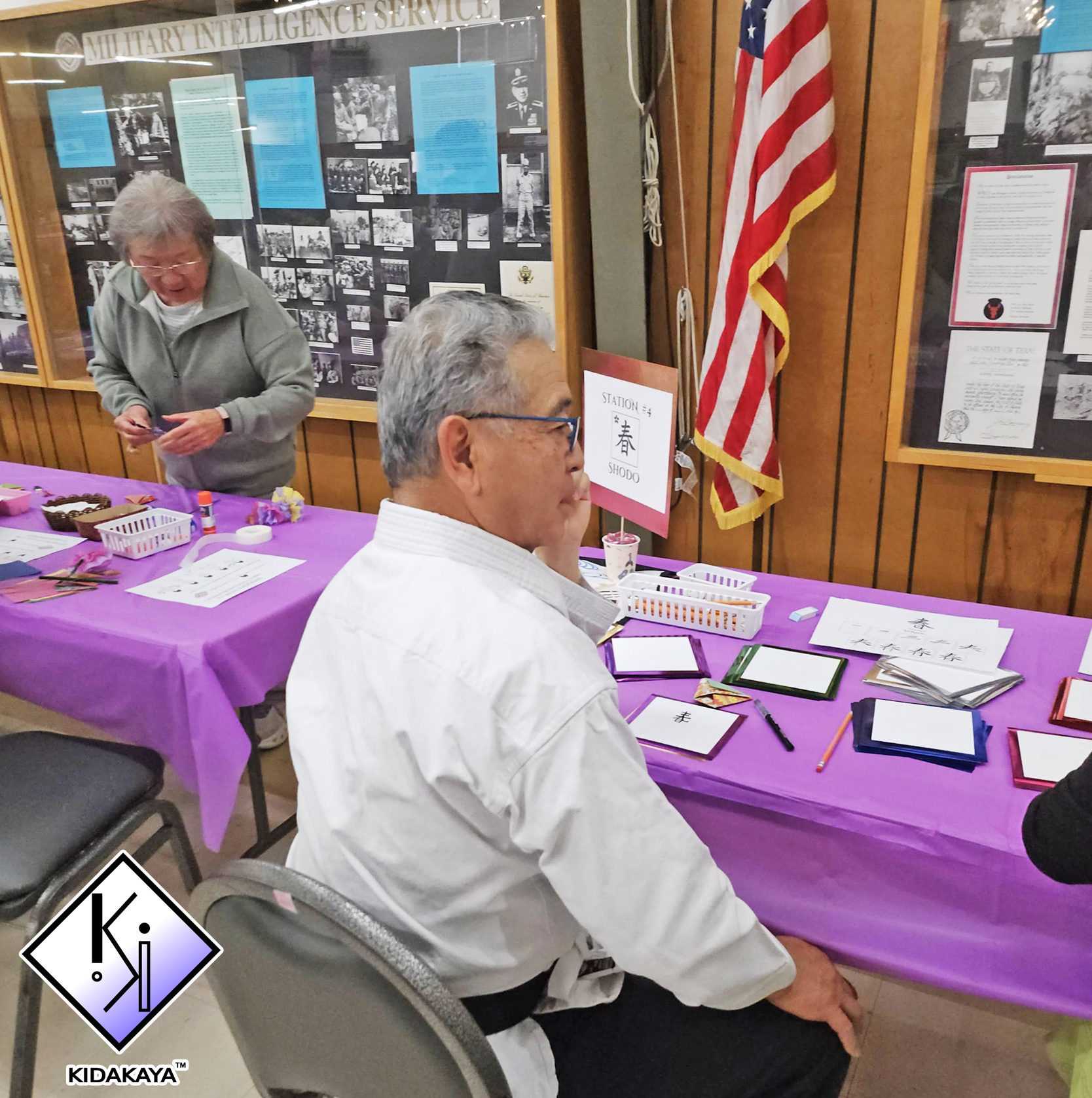 An elderly man in a white shirt sitting at a purple table with paper and markers, looking to his side. A woman standing behind the table, wearing a gray jacket, is smiling and holding some papers. The table has various crafts materials. Behind them i