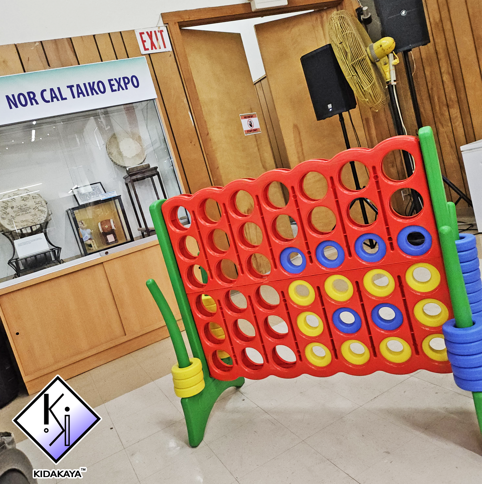 Indoor setting with a large, colorful Connect Four game in the foreground. A wooden cabinet with glass display case containing various artifacts is visible in the background. A sign reading 'NOR CAL TAIKO EXPO' is on the wall, along with an 'EXIT' si