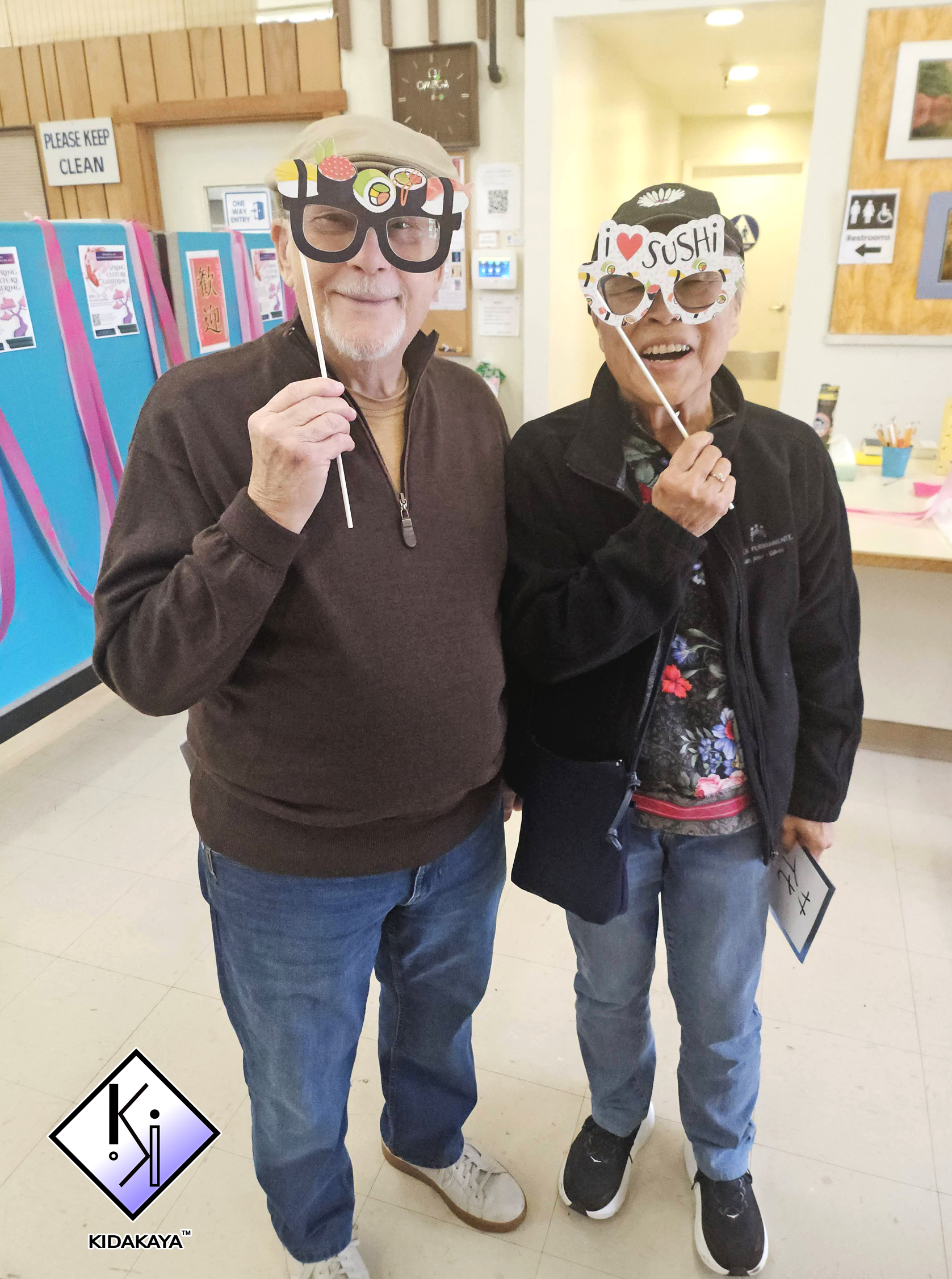 Two older adults standing indoors, wearing novelty sushi-themed masks, smiling and holding their masks on sticks.
