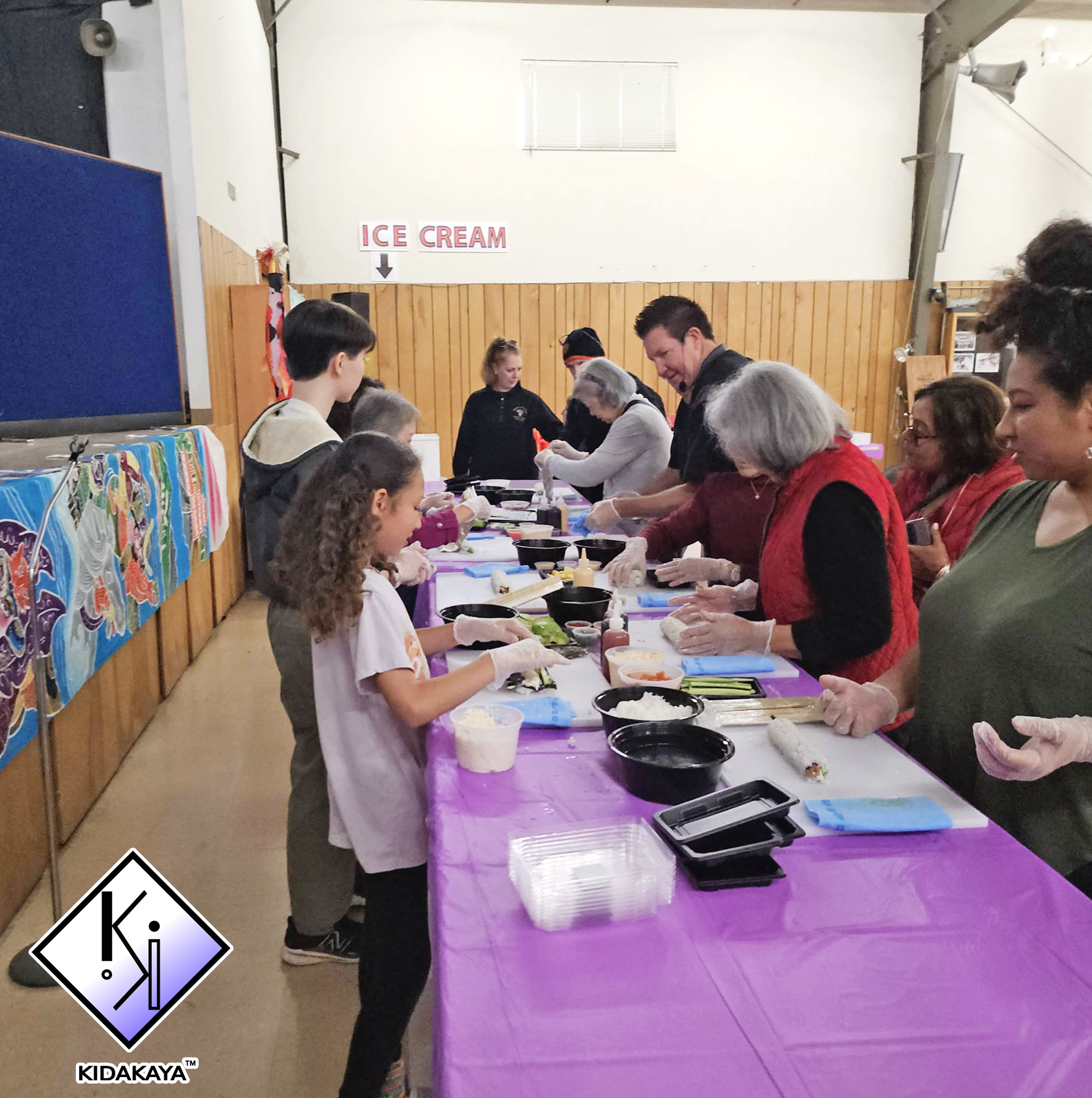 A group of people, including children and adults, are gathered around a long table covered with a purple tablecloth, making sushi. There are various bowls of ingredients, sushi mats, and other supplies on the table. In the background, a sign reads "I